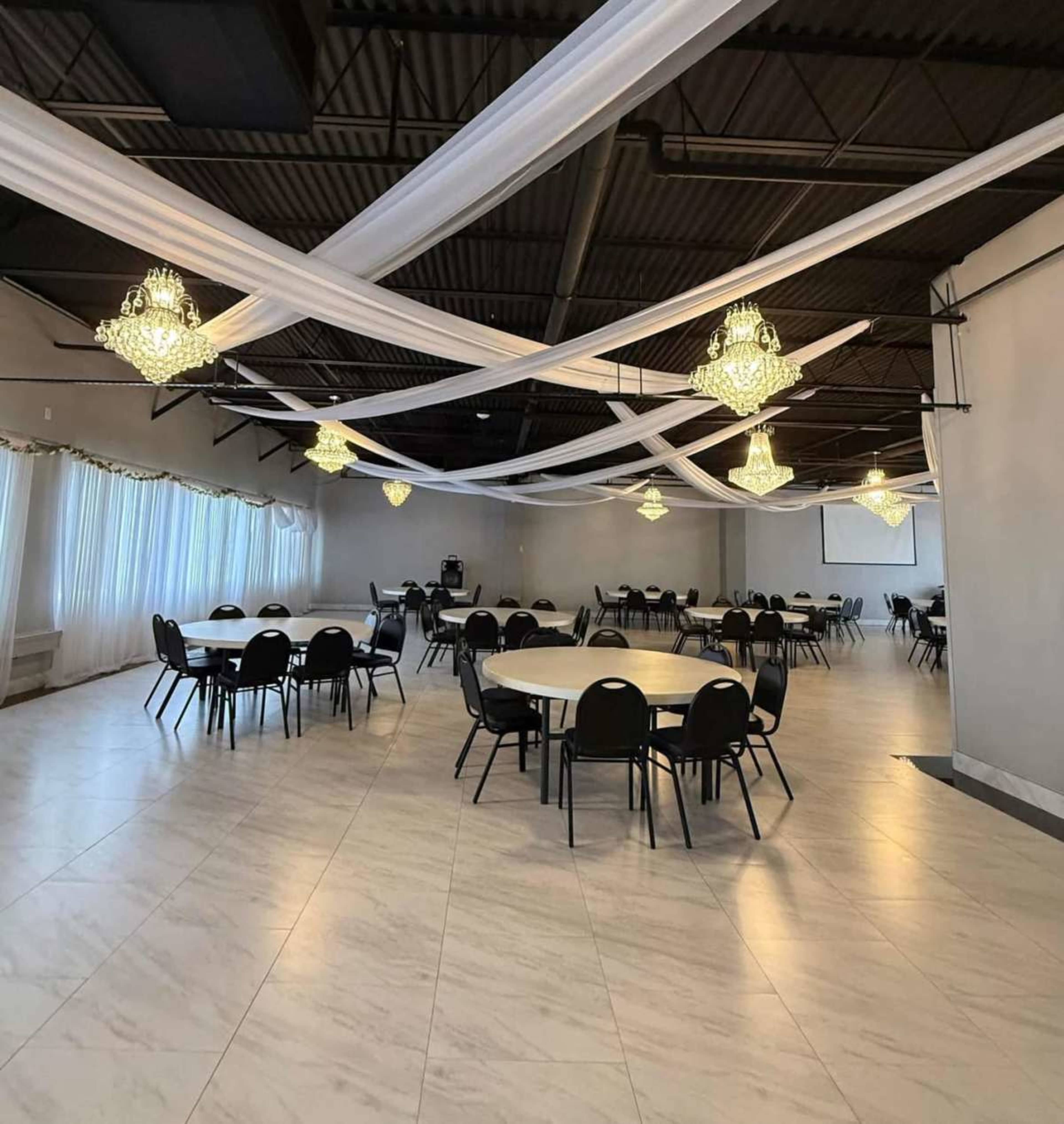 The image shows a spacious event hall with round tables and black chairs, adorned with chandeliers and draped white fabric across the ceiling.