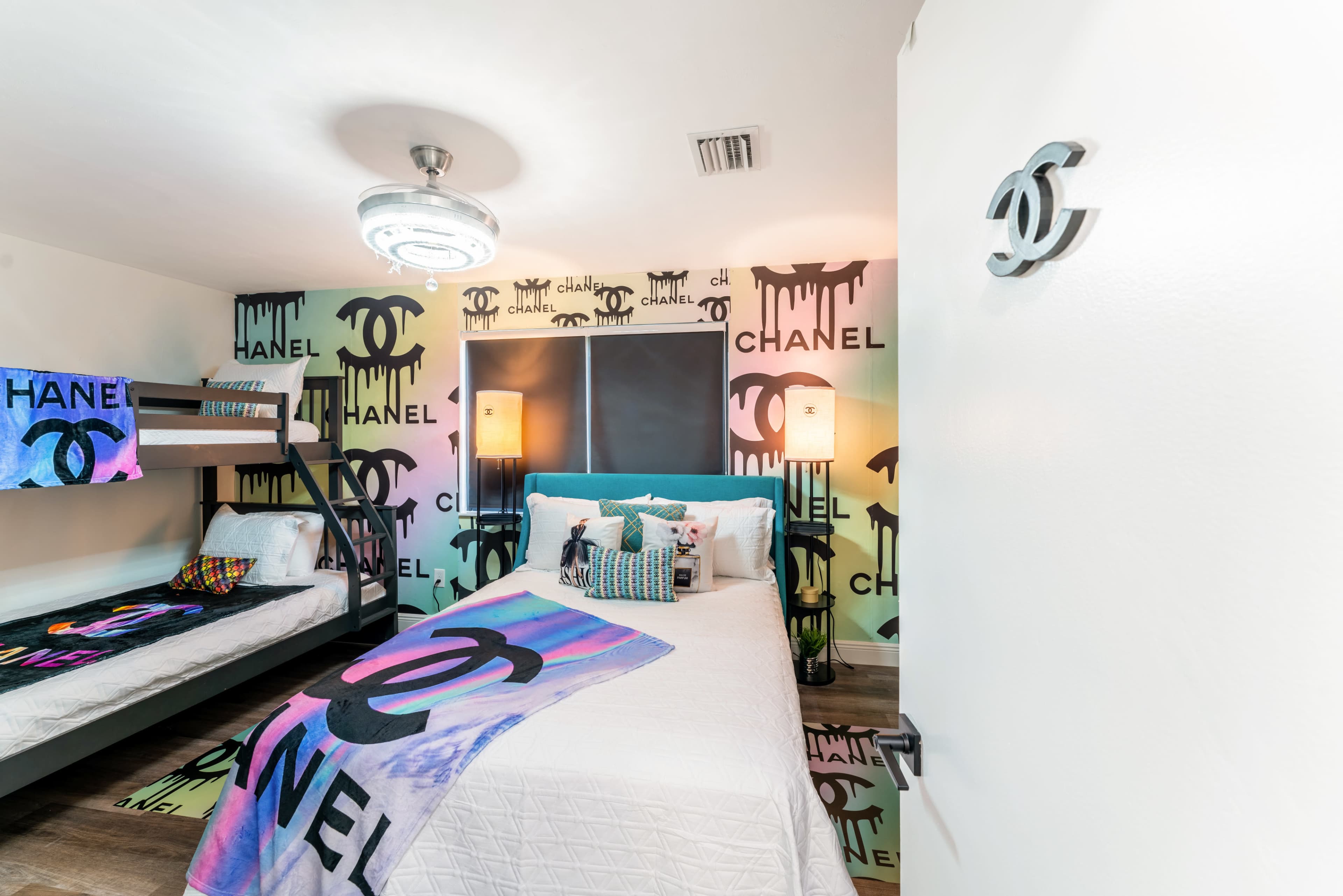 A brightly decorated bedroom featuring a bunk bed, a single bed, and walls adorned with colorful Chanel logos.