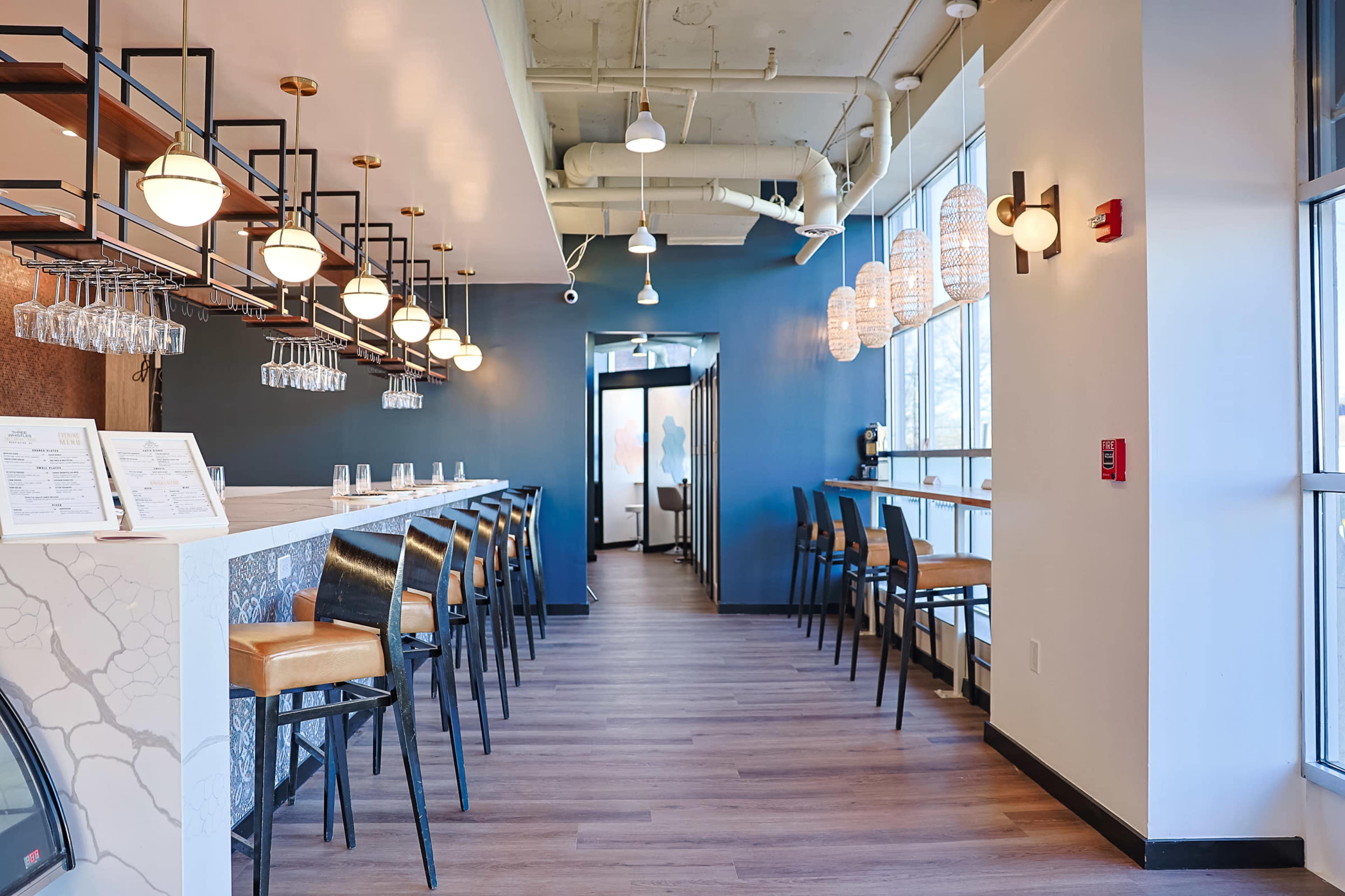 The image shows a modern restaurant interior featuring a long bar with pendant lights, stools, and a row of hanging glassware along one side, leading to a hallway.