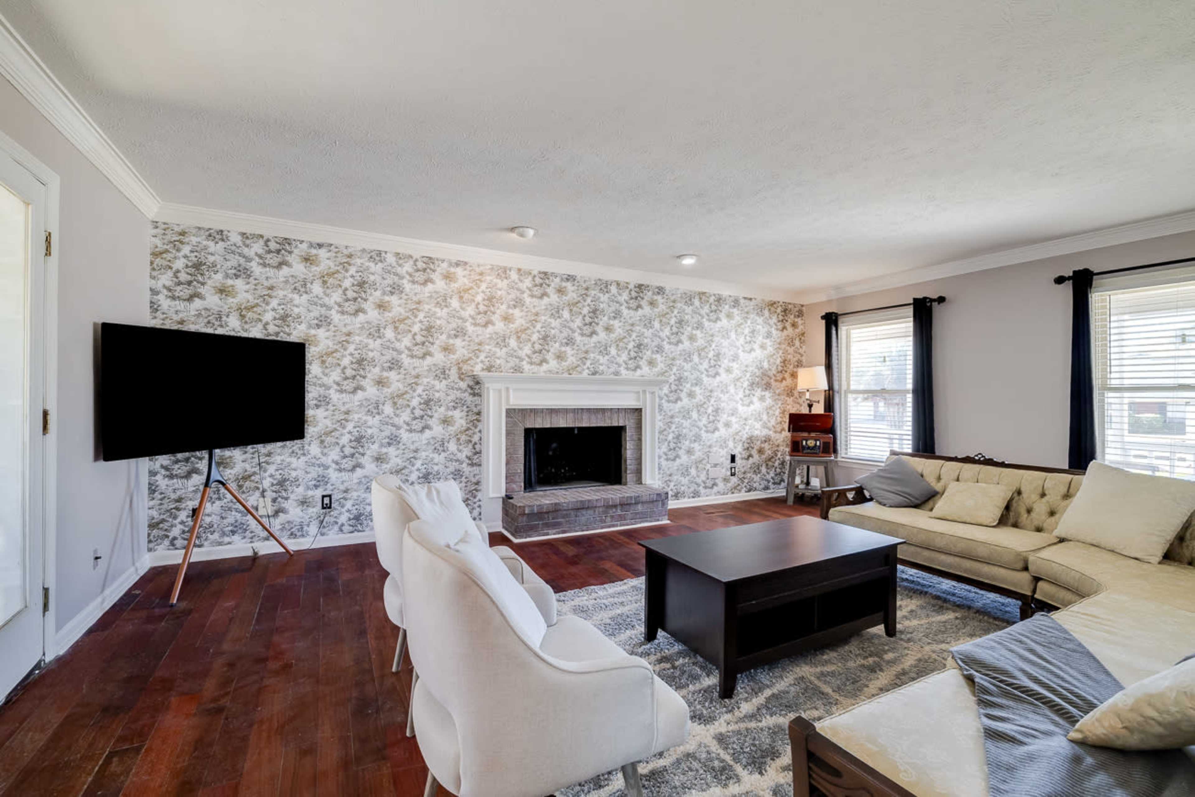 The living room features a fireplace against floral wallpaper, a flat-screen TV on a stand, a beige sectional sofa, and a coffee table on a rug.