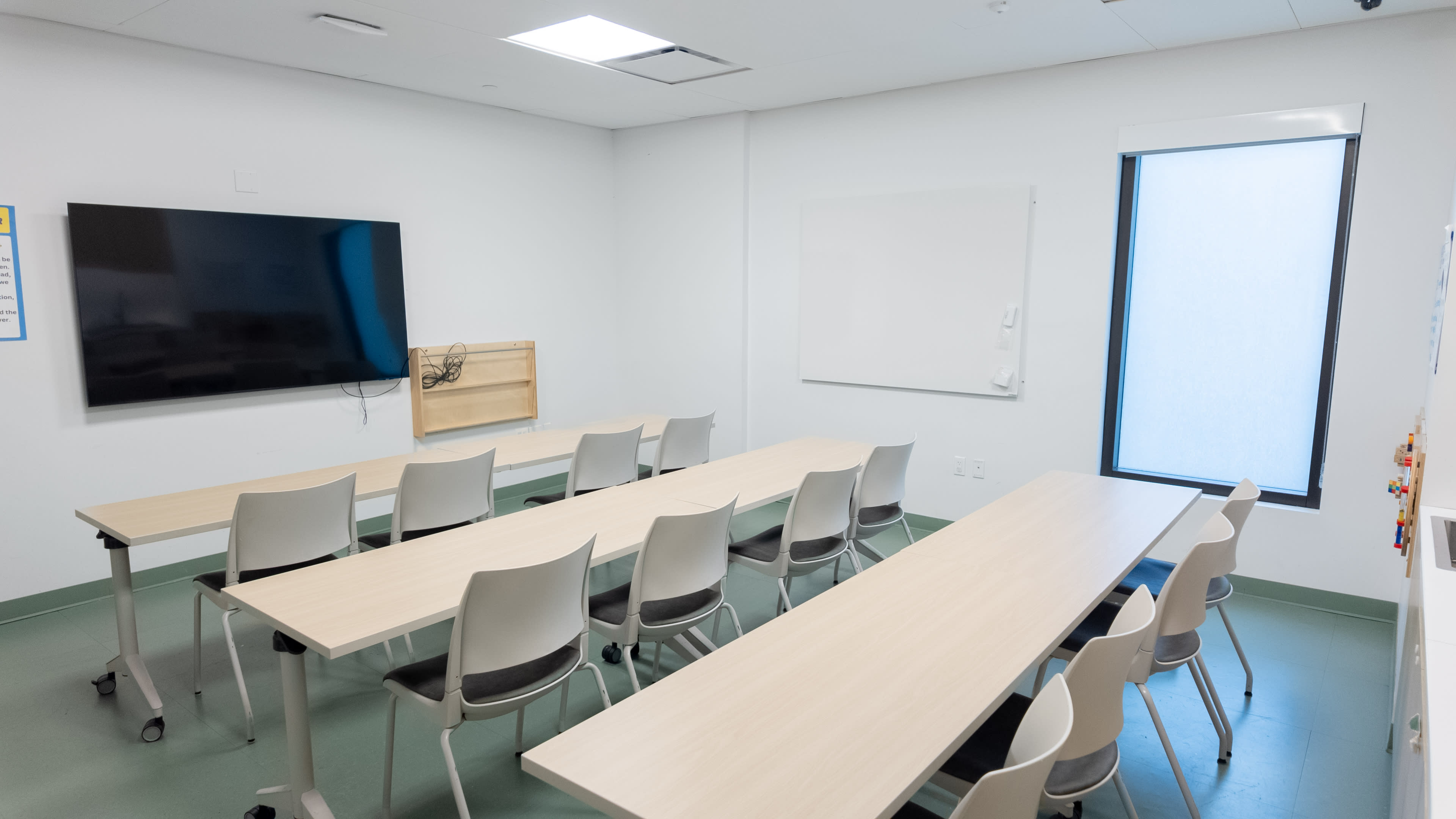 The image shows a classroom set up with multiple tables and chairs, a large wall-mounted television, and a whiteboard.