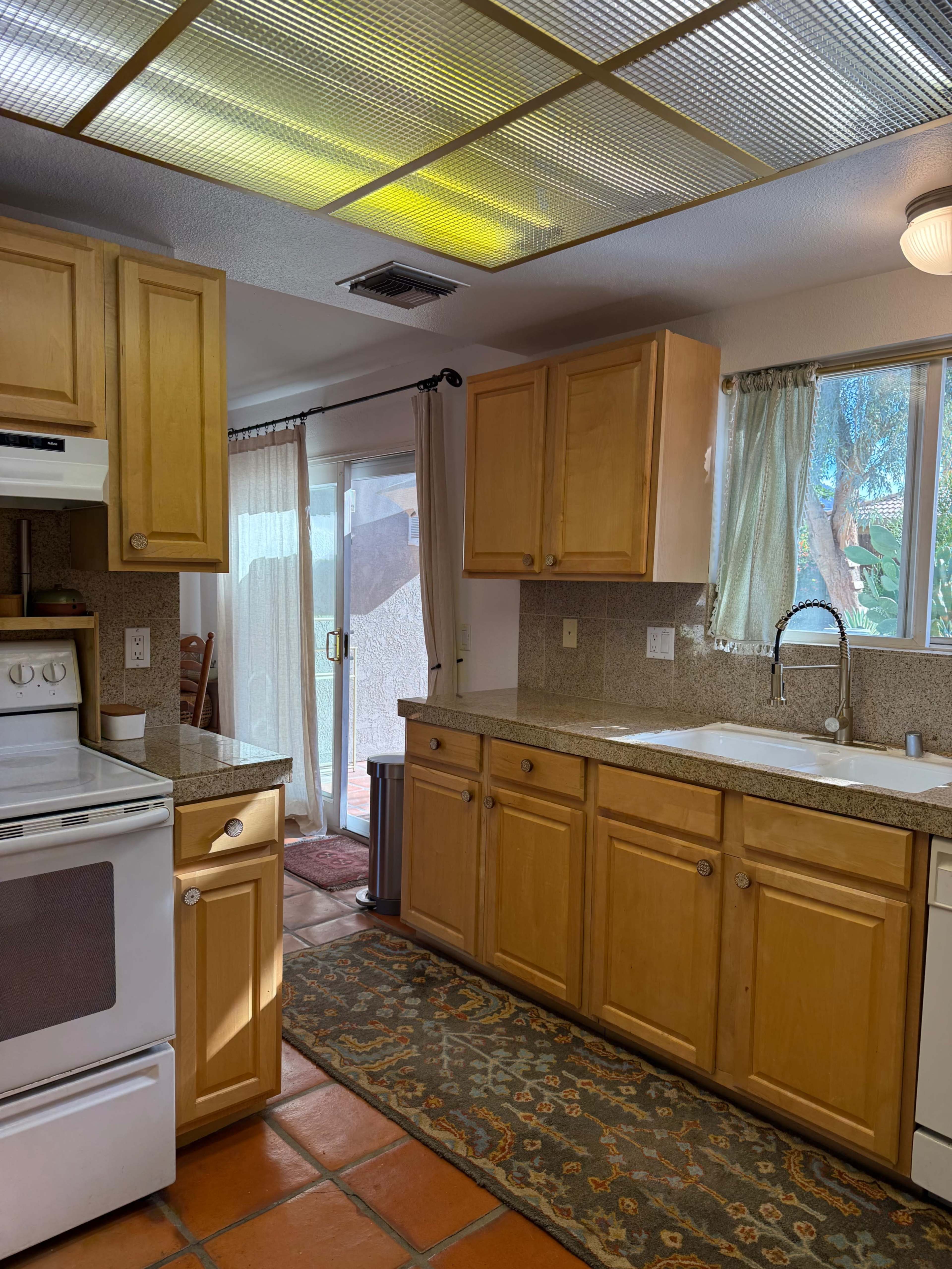 The image shows a kitchen with wooden cabinets, a sink under a window, and a stove, featuring tiled flooring and a door leading outside.