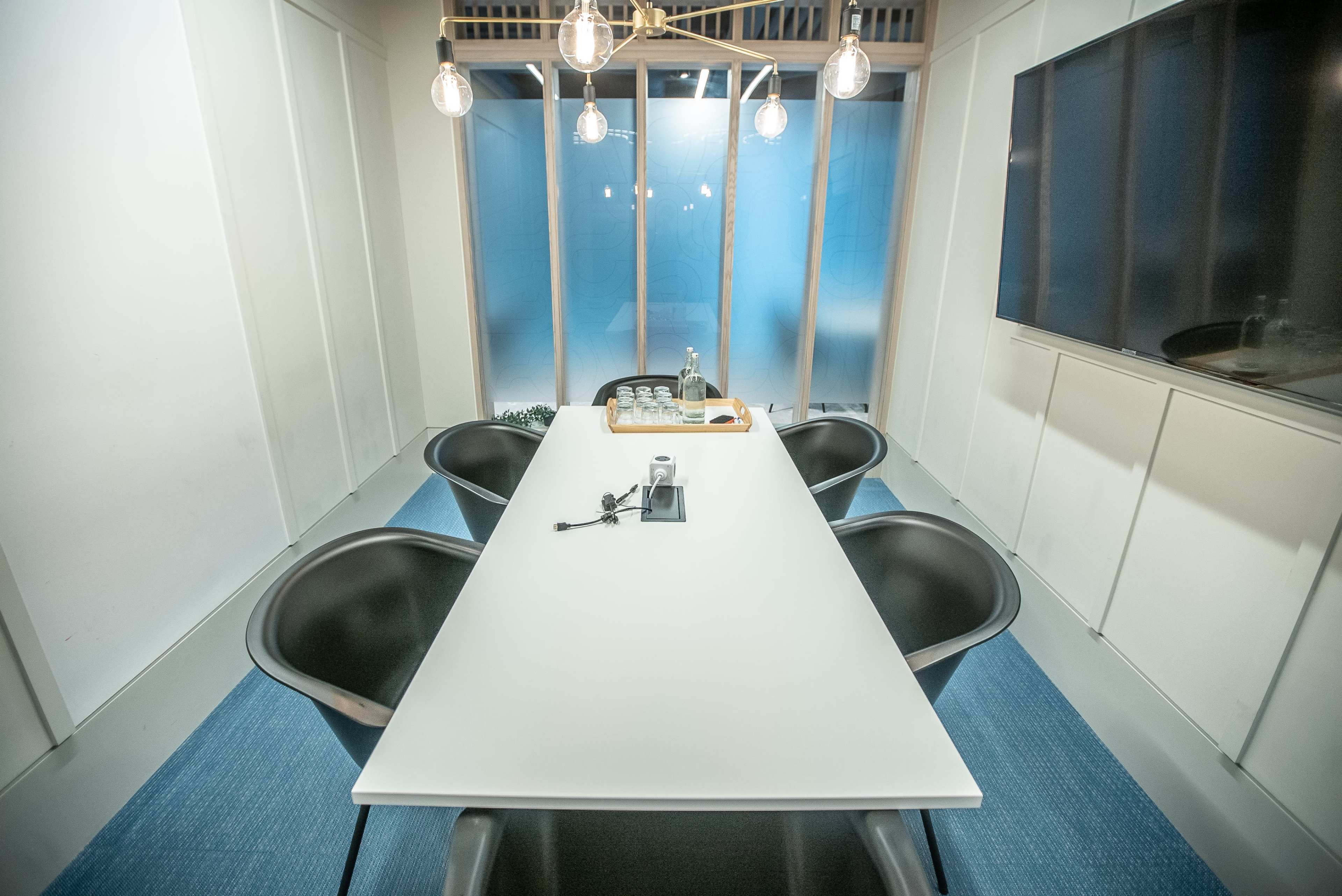 A modern conference room features a long white table surrounded by black chairs, with a large screen on one wall and glass windows allowing natural light.