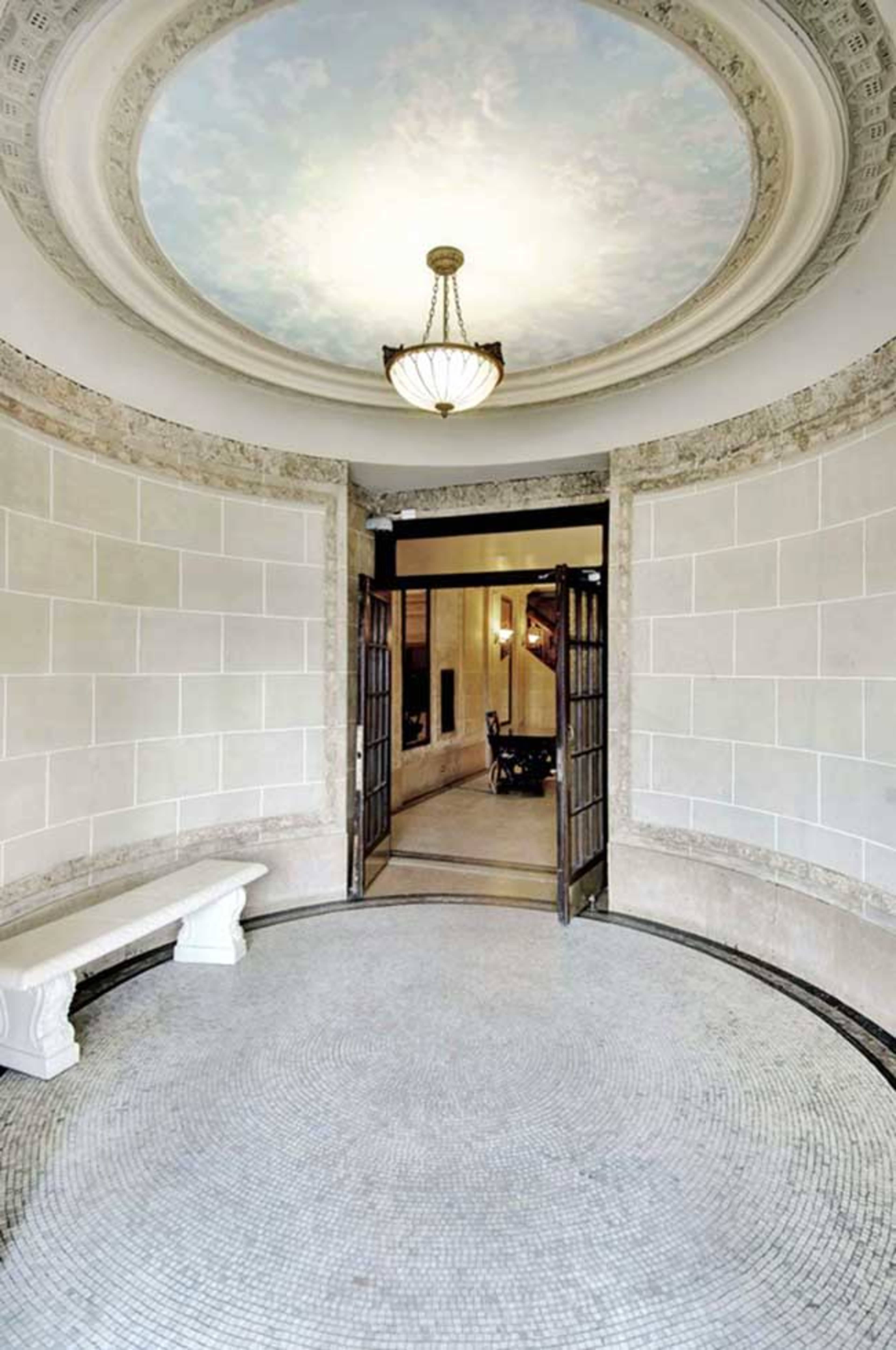 The image shows a circular entryway with a decorative ceiling and double doors leading to another room, featuring a curved stone bench and tiled floor.