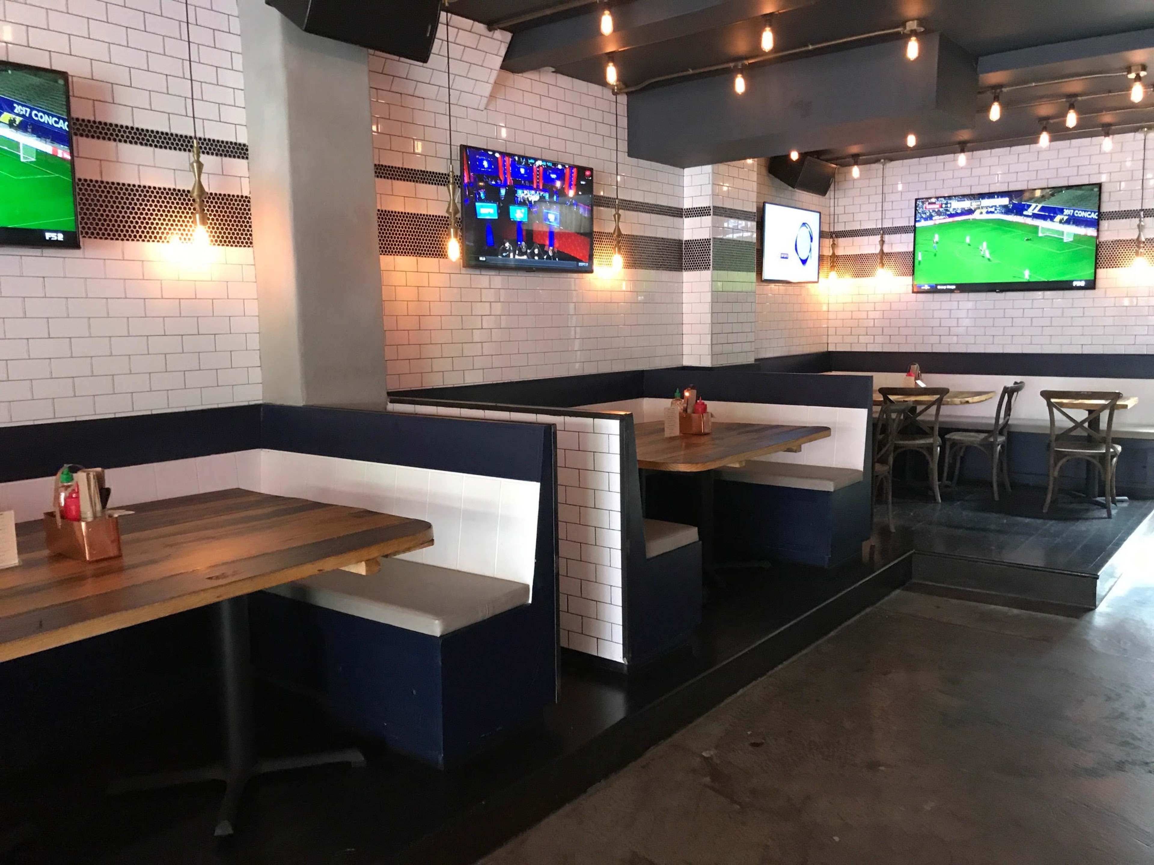 A modern restaurant interior with wooden tables, white tiled walls, and several televisions displaying sports.