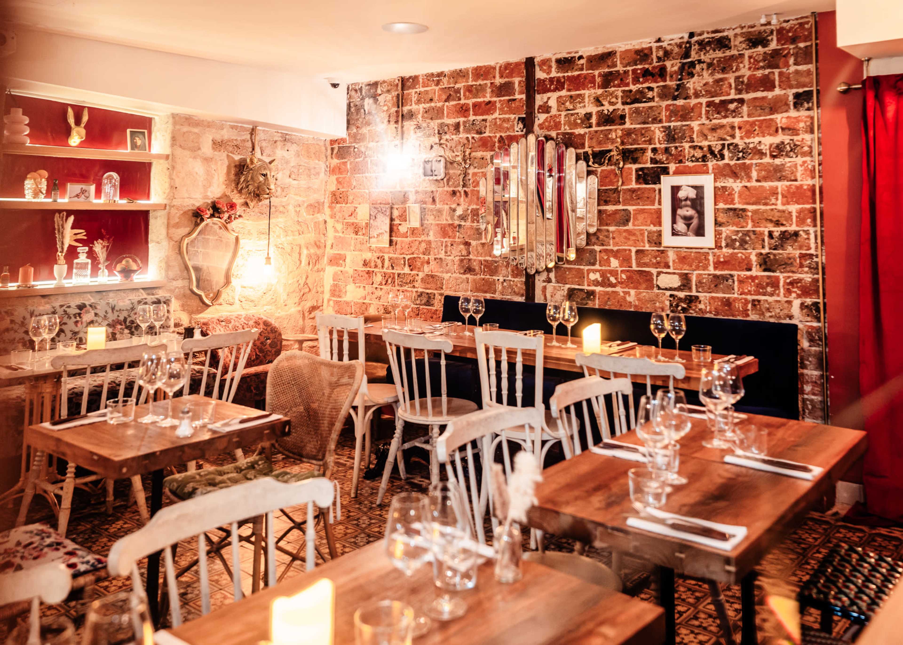An intimate restaurant featuring wooden tables set with glassware, a brick wall adorned with mirrors, and warm lighting creating a cozy atmosphere.