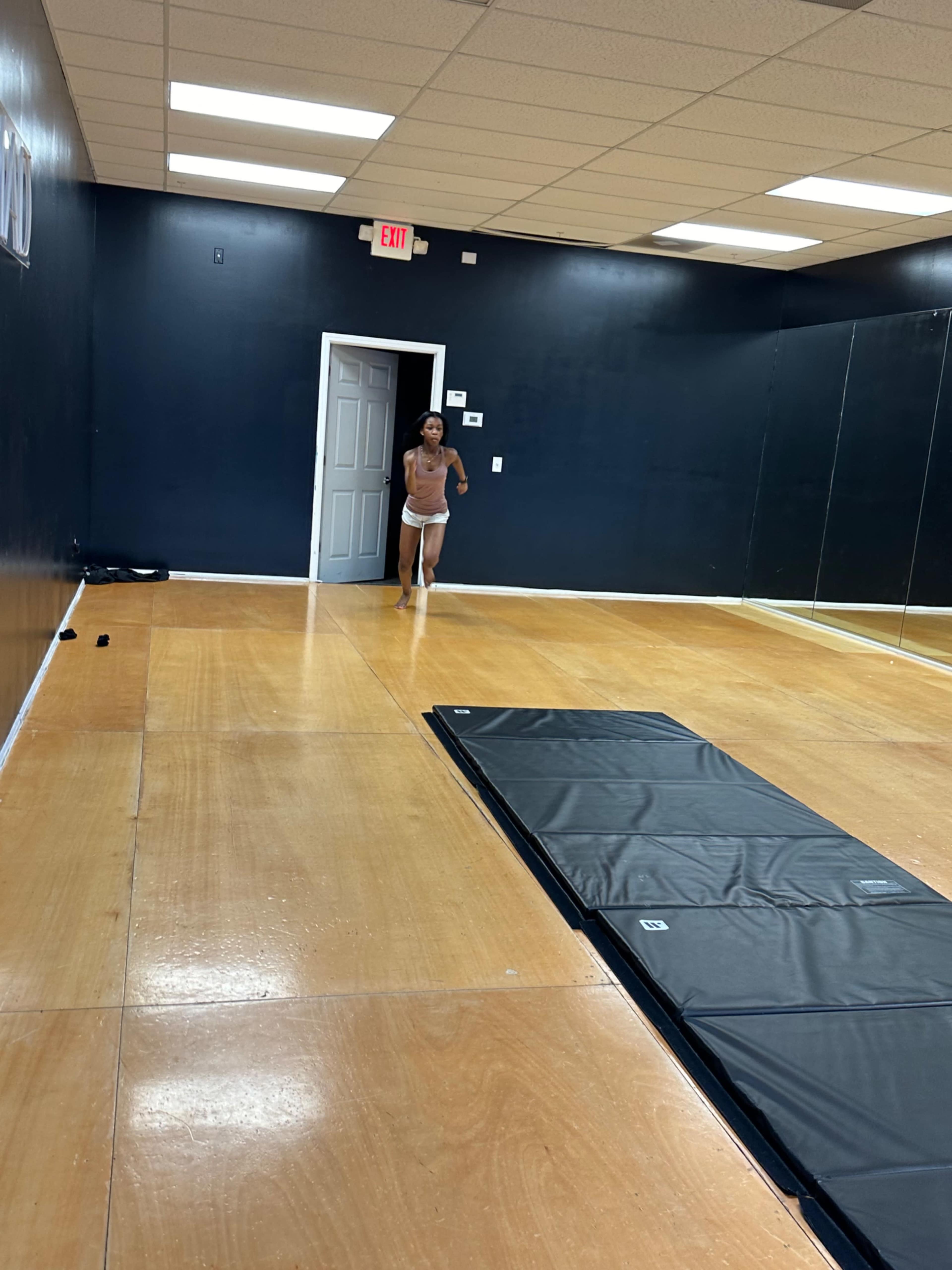 A person is walking into a dance studio with a mirrored wall and a padded mat on the floor.