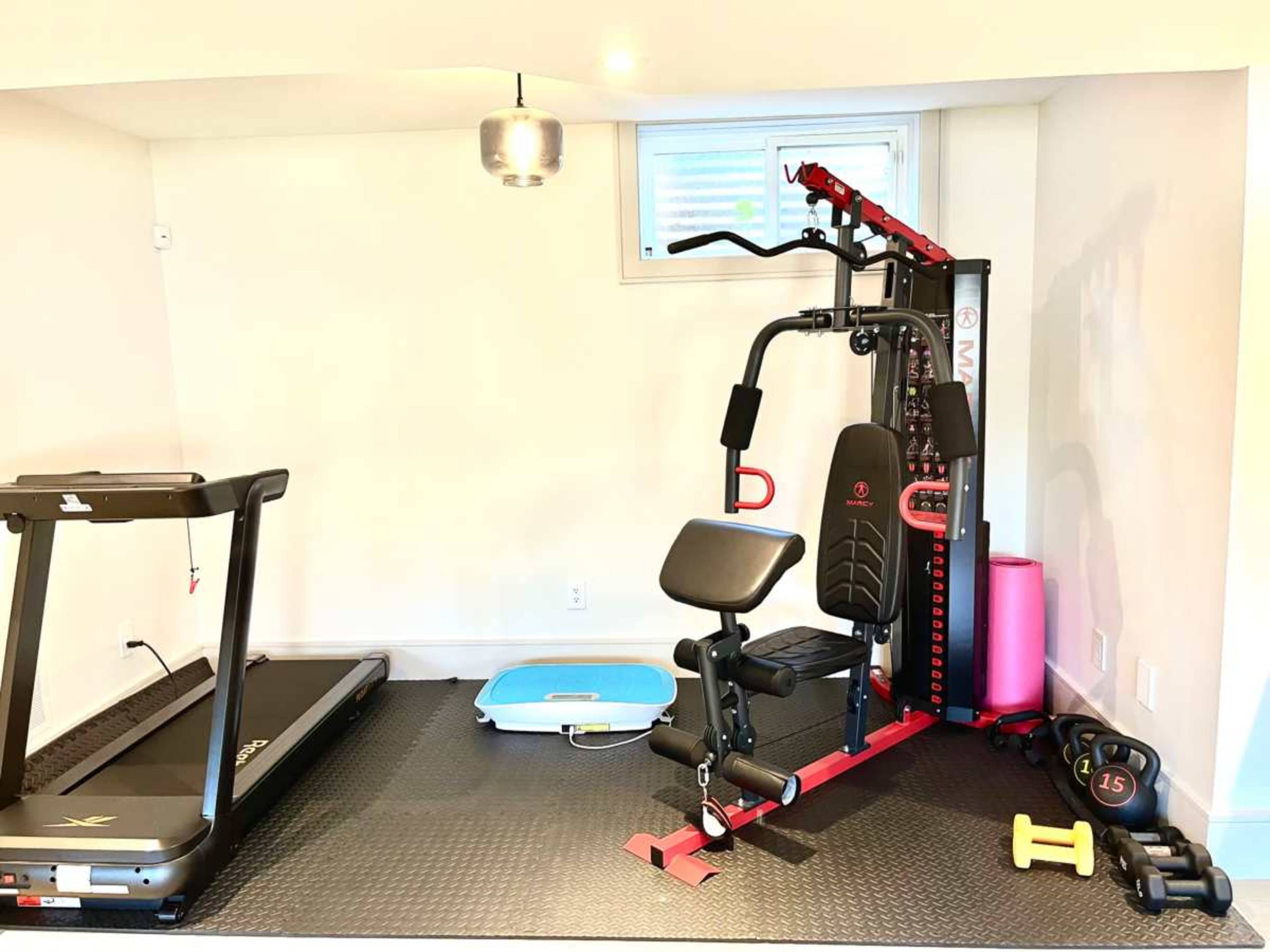 The image shows a gym setup featuring a treadmill, a multi-gym machine, a blue exercise mat, and various weights on a rubber floor mat.