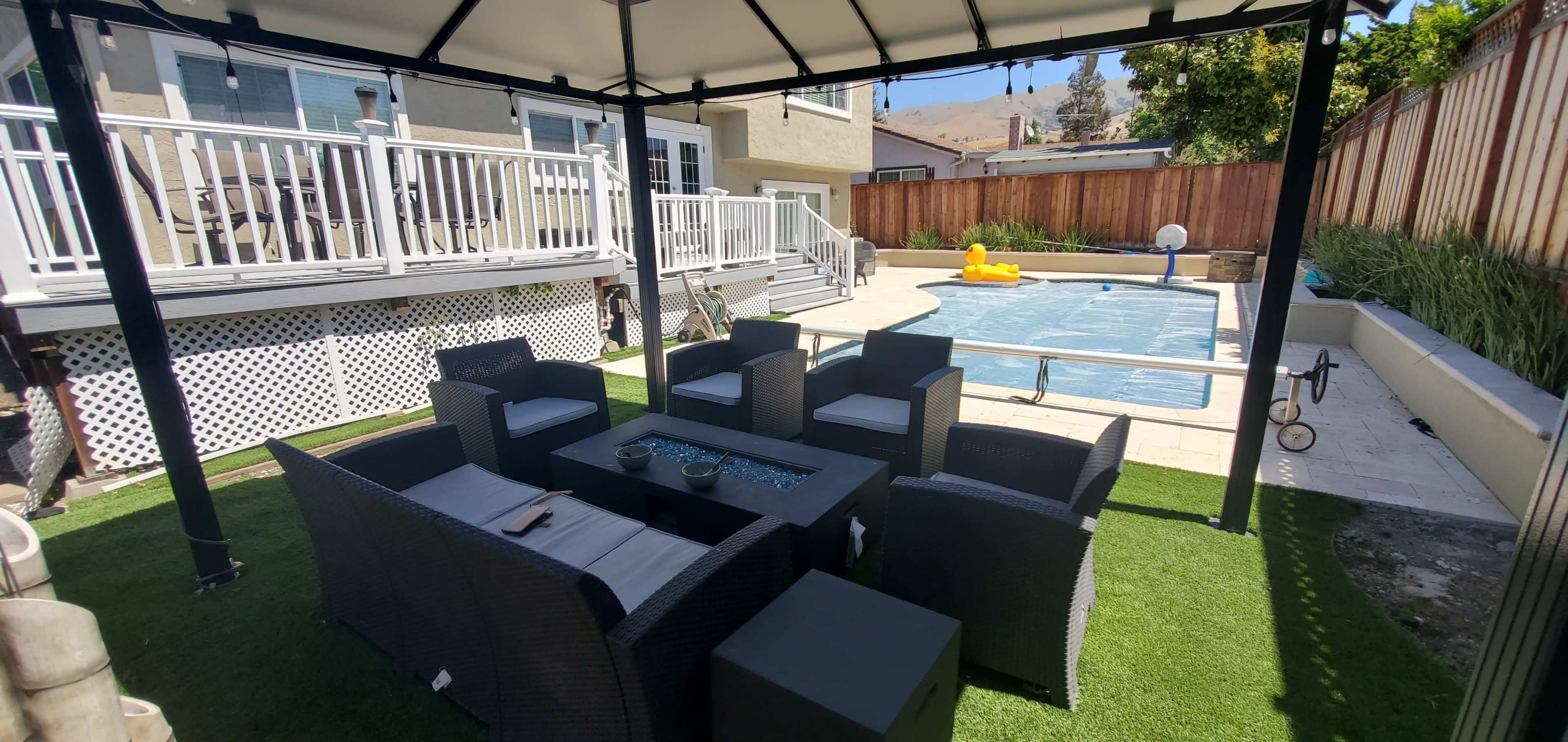 A shaded outdoor seating area with black wicker furniture surrounds a glass table, overlooking a pool with a yellow float.