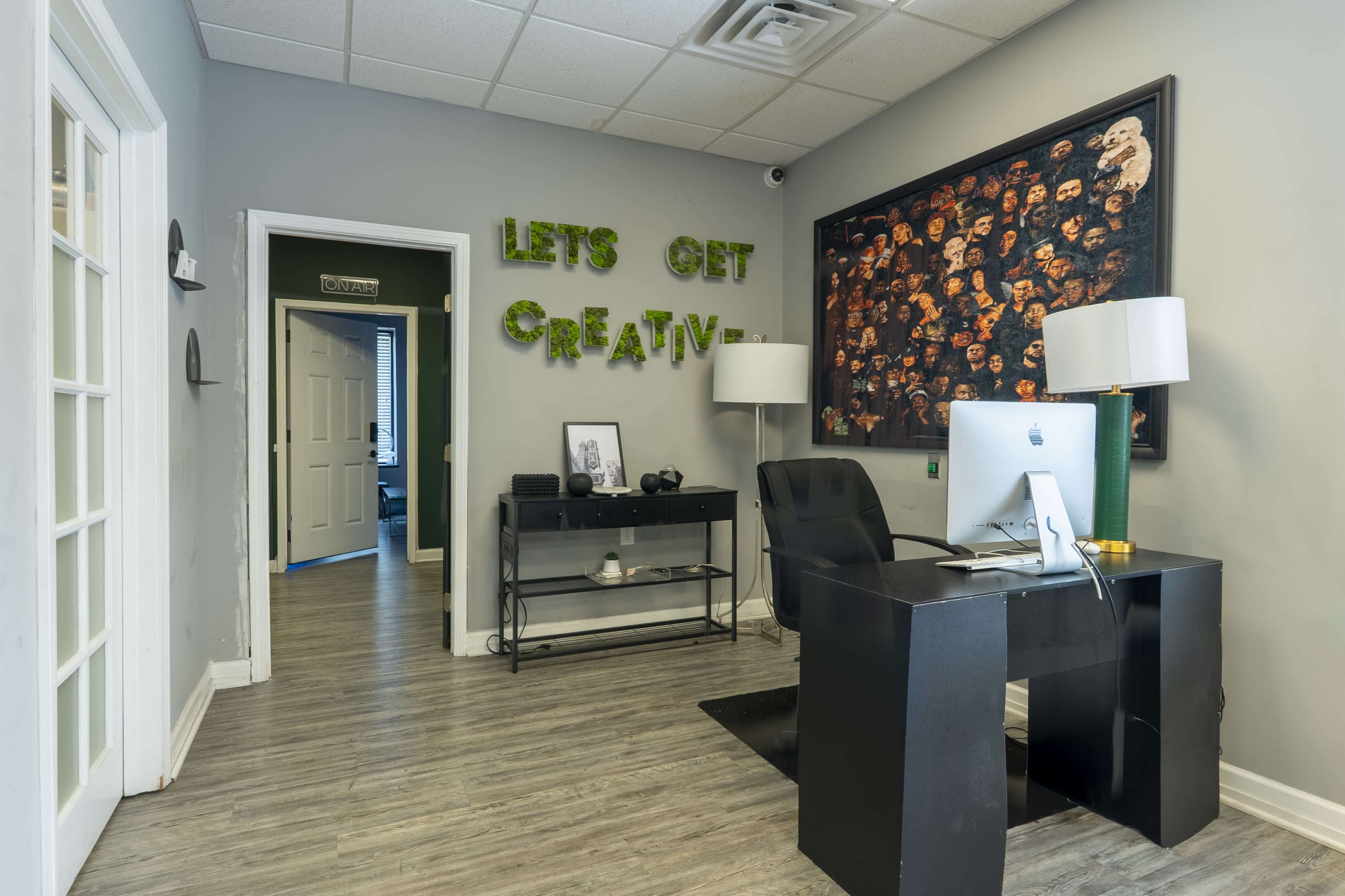 The image shows an office space featuring a black desk with a computer, a wall adorned with a large framed artwork, and the phrase "LET'S GET CREATIVE" in green letters on the wall.