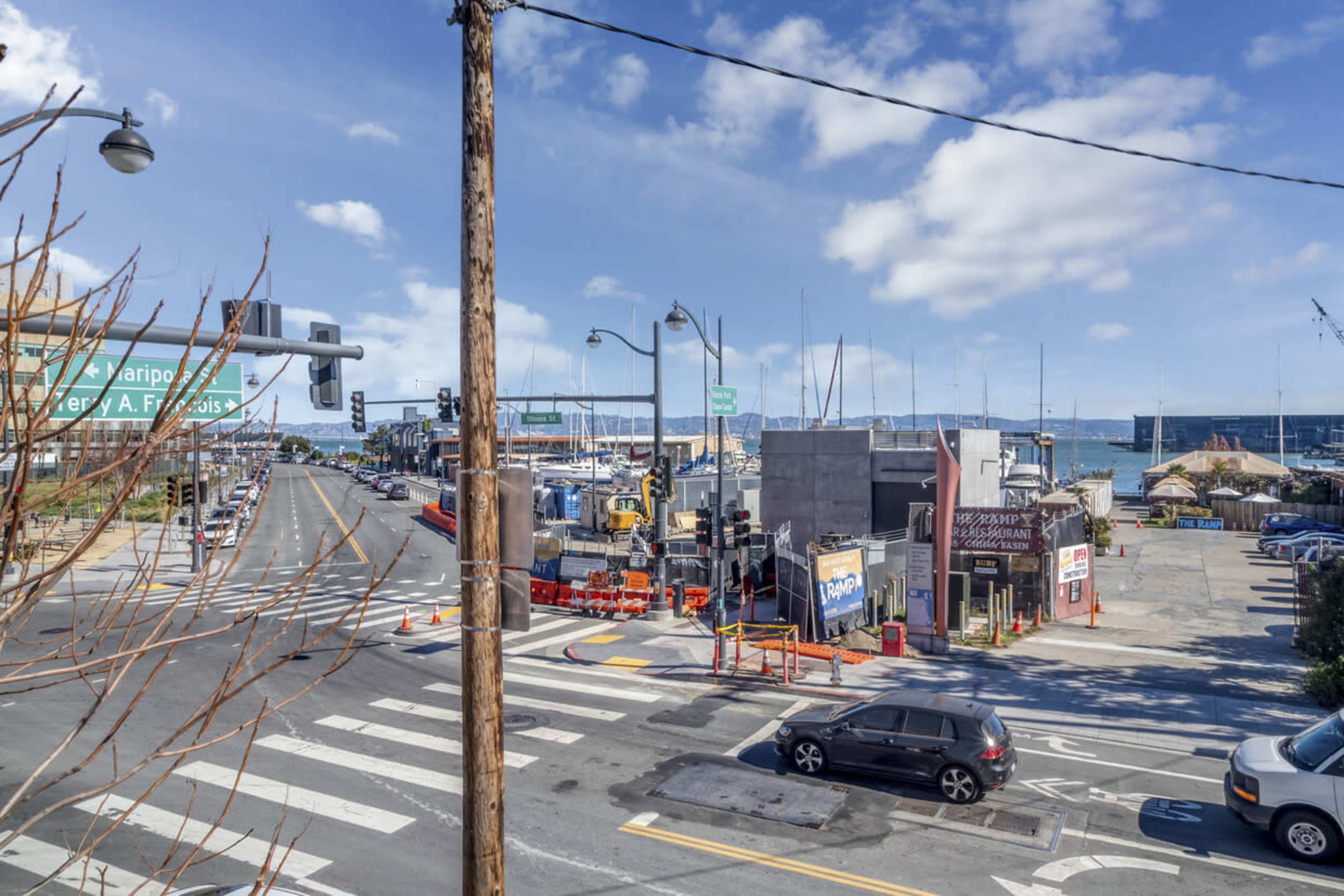 A busy street intersection near a marina, with parked cars, construction equipment, and various businesses visible.