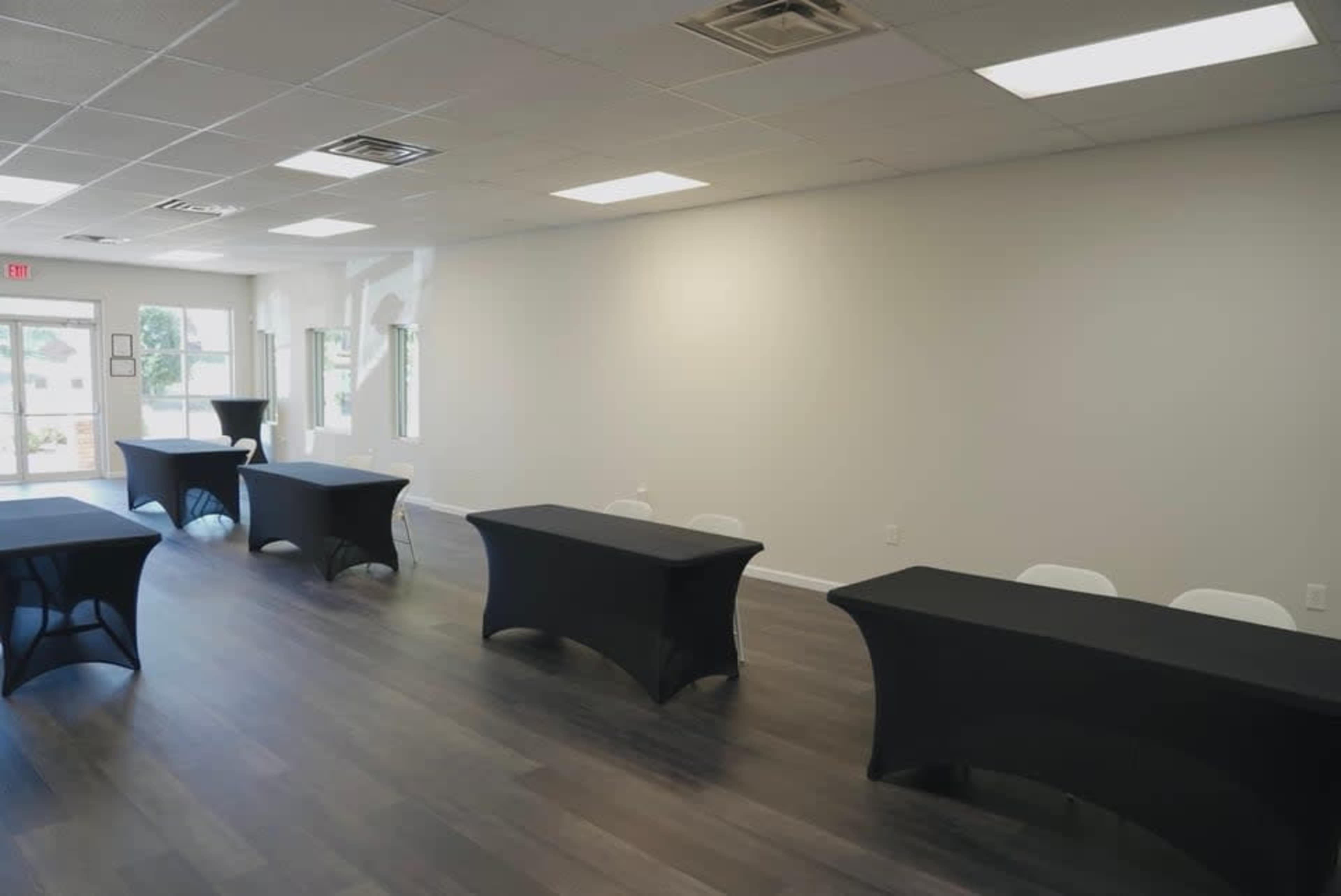 The image shows a room with a polished wooden floor and several black-covered tables arranged in a row, with large windows and natural light coming from the entrance.
