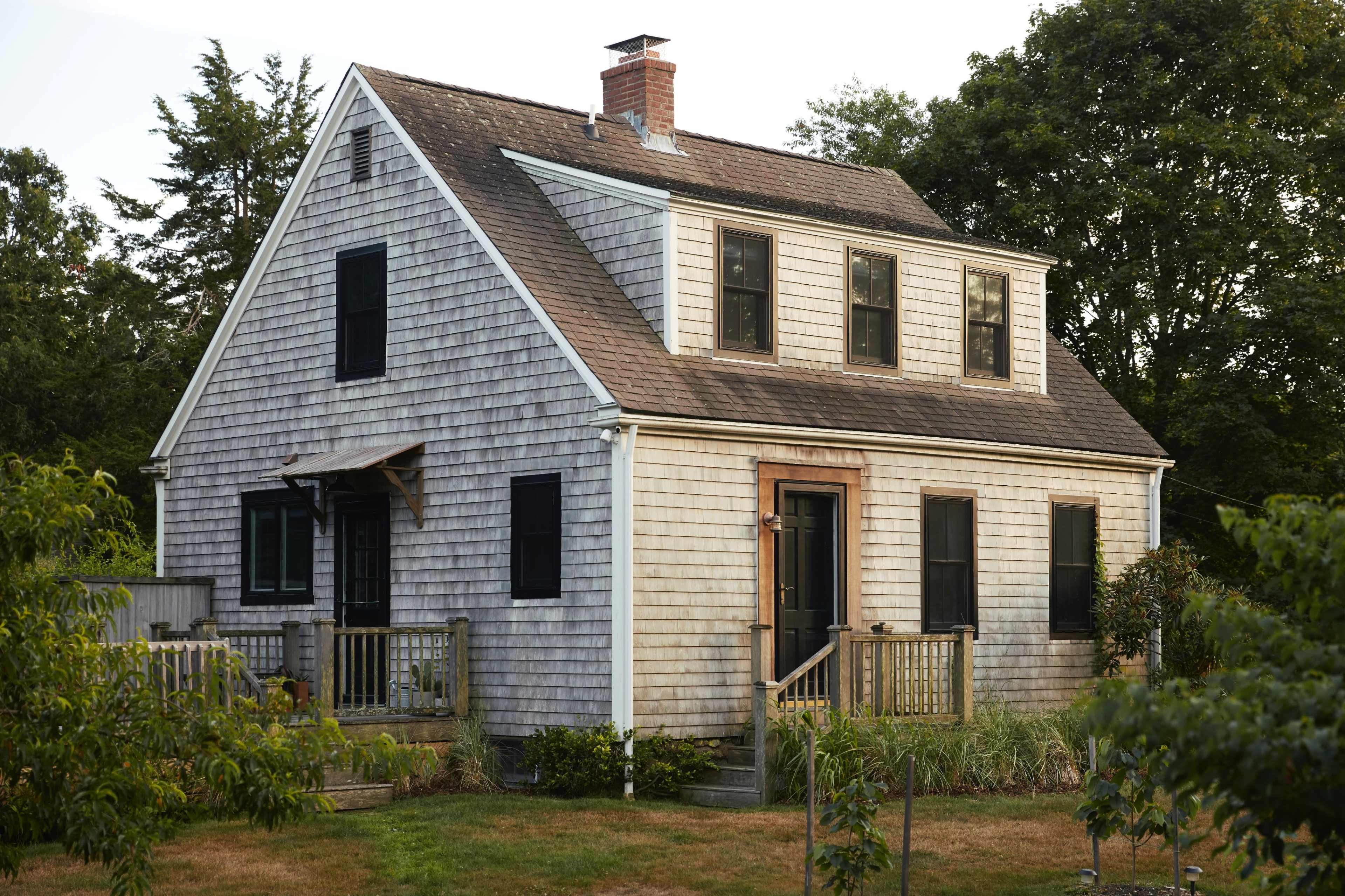 A weathered, two-story house with a steep roof and wooden shingles is surrounded by a grassy area and trees.