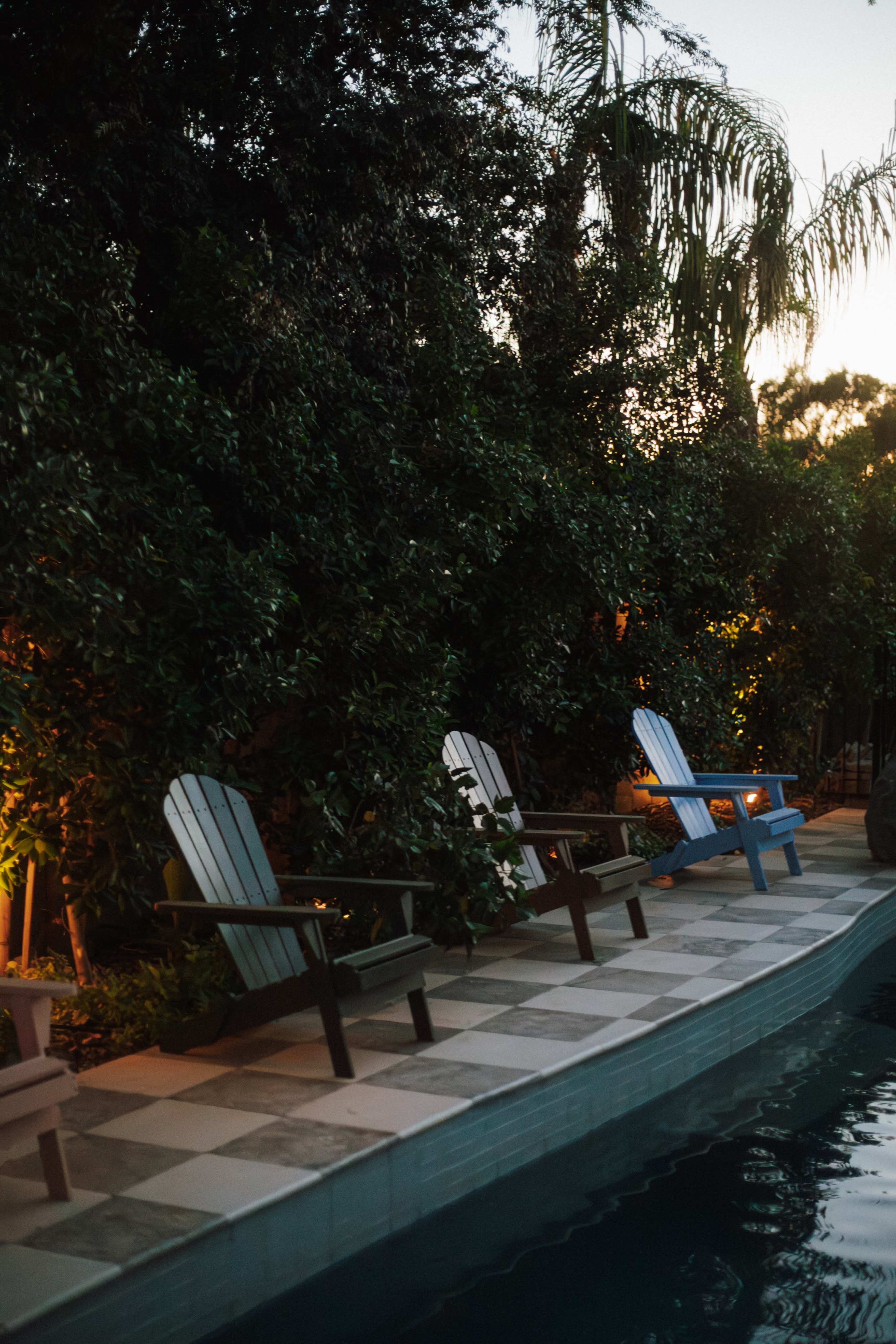 The image shows a tranquil poolside area with several adirondack chairs arranged along the edge of the water, surrounded by lush greenery and soft lighting.