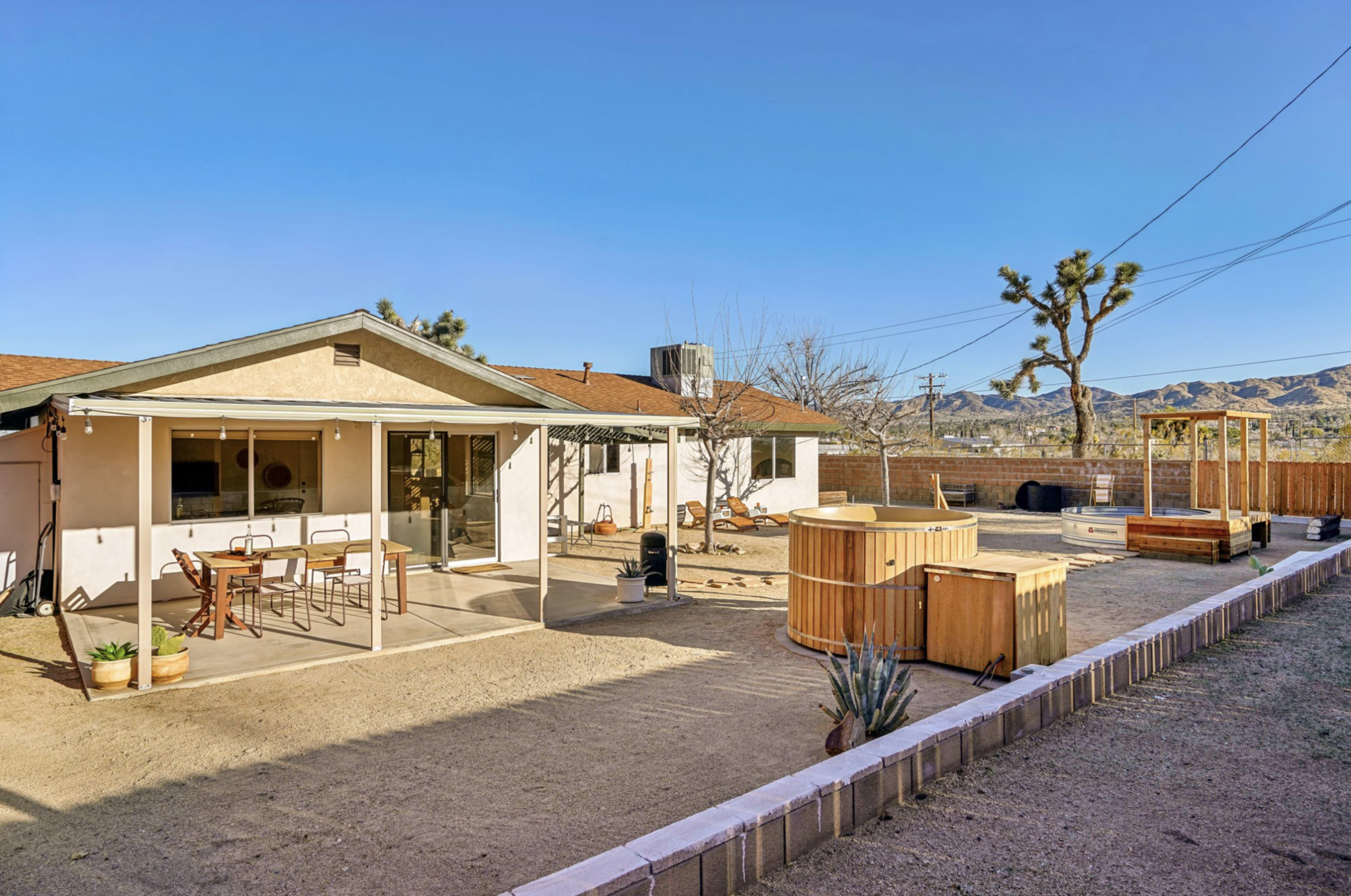 A desert backyard features a house with a patio, a wooden hot tub, and sparse vegetation against a clear blue sky.