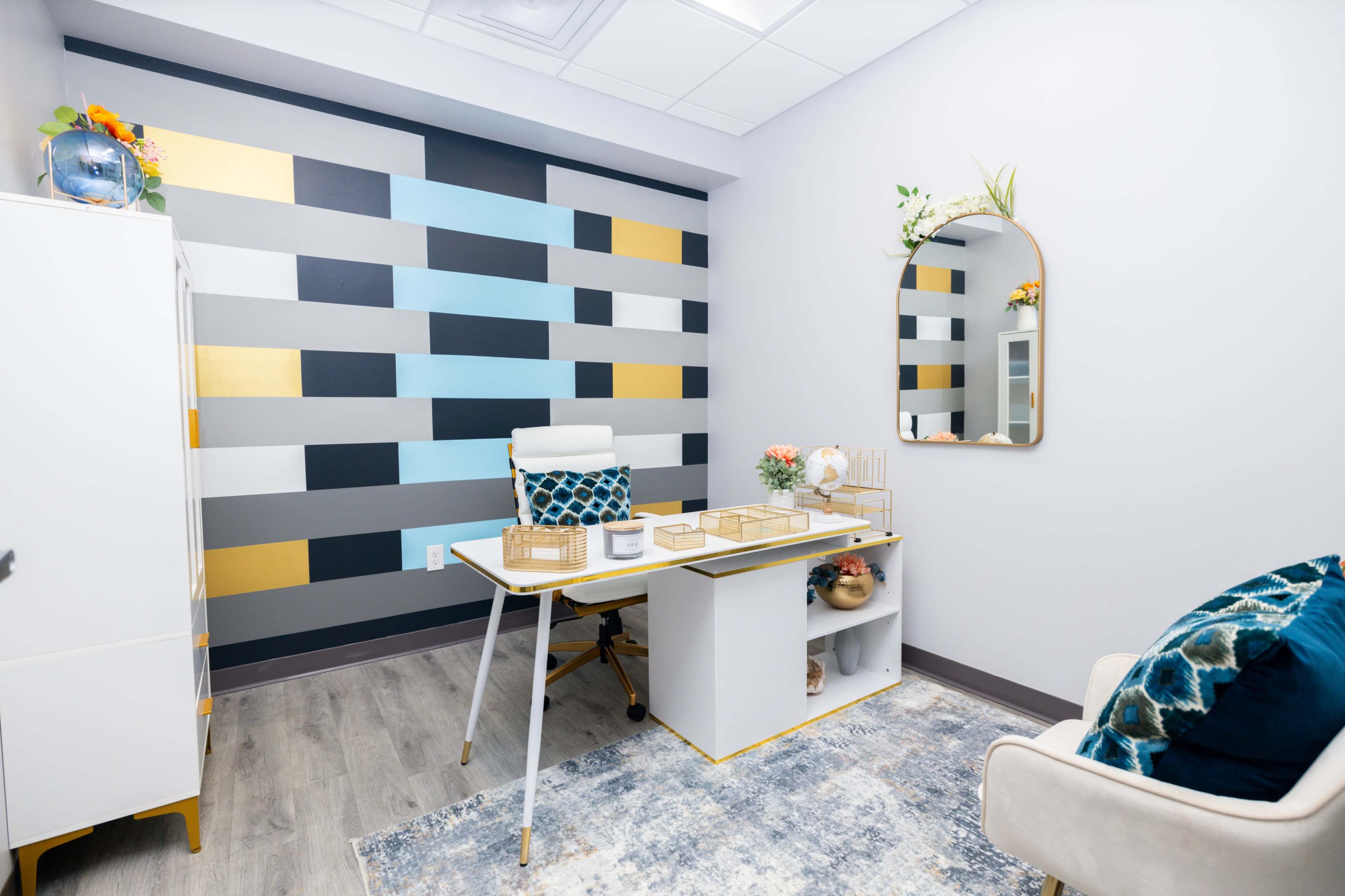 The image shows a modern office space featuring a white desk with organized decorative elements, a mirror, a chair, and a striped wall in various colors.