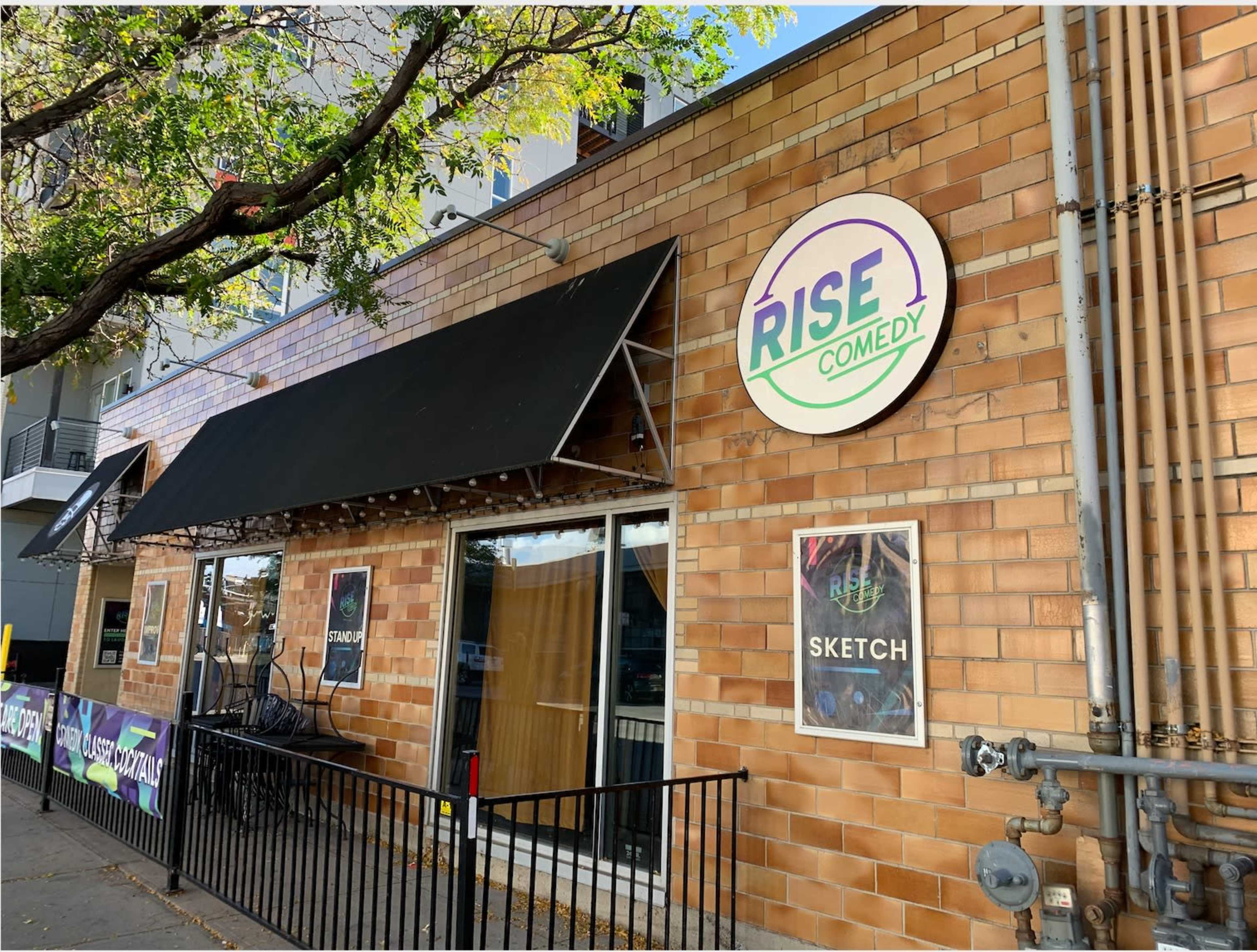 A brick building with a black awning and a circular sign reading "RISE COMEDY" is visible, along with posters displayed at the entrance.