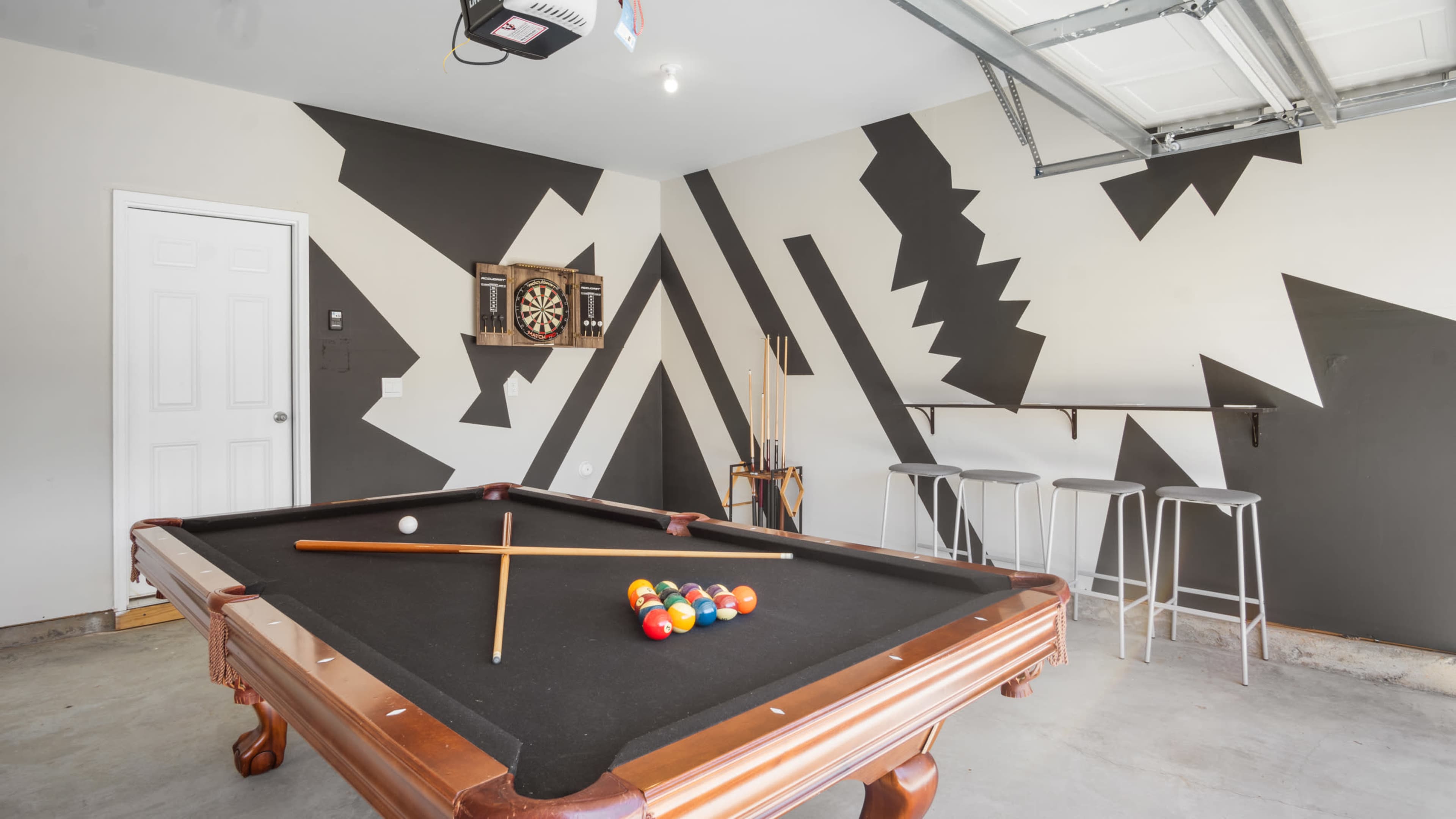 A garage with a billiard table, a dartboard on the wall, and modern bar stools along one side.