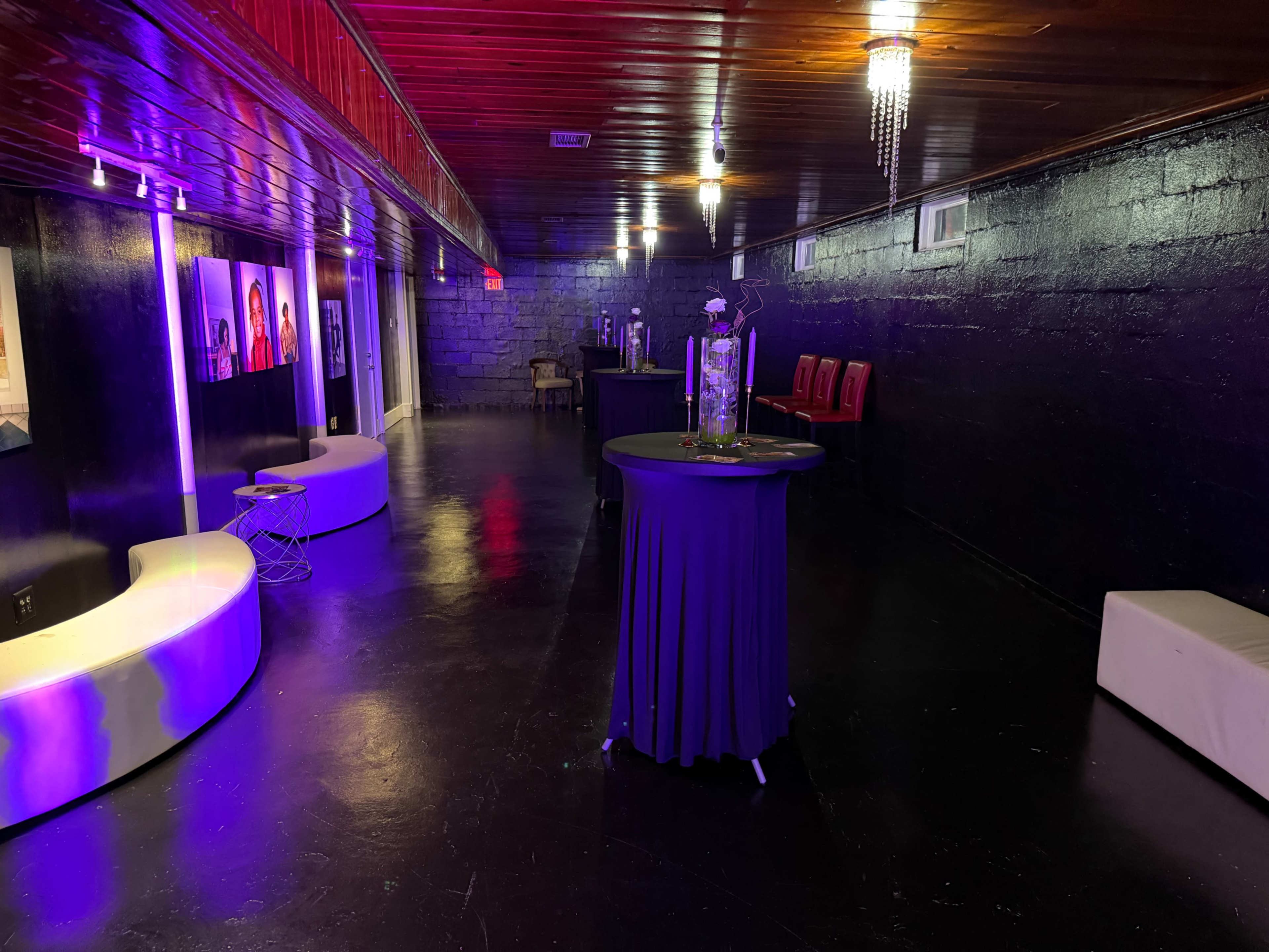 The image shows a dimly lit event space with glossy black floors, featuring a few tables with tablecloths, seating arrangements against the walls, and decorative lighting.