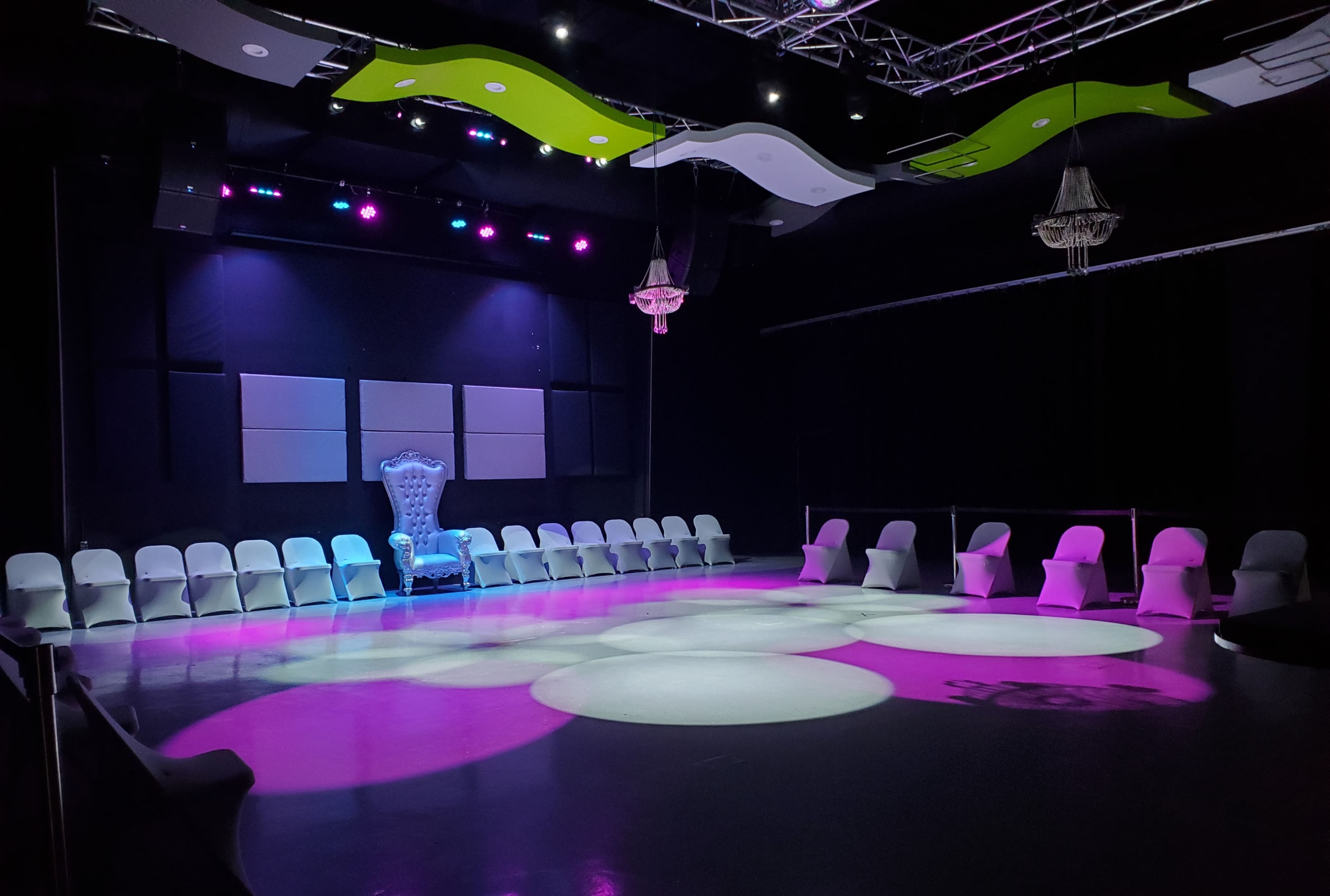 A dimly lit event space with arranged white chairs surrounding a circular area, featuring a large blue throne under purple and green lighting.
