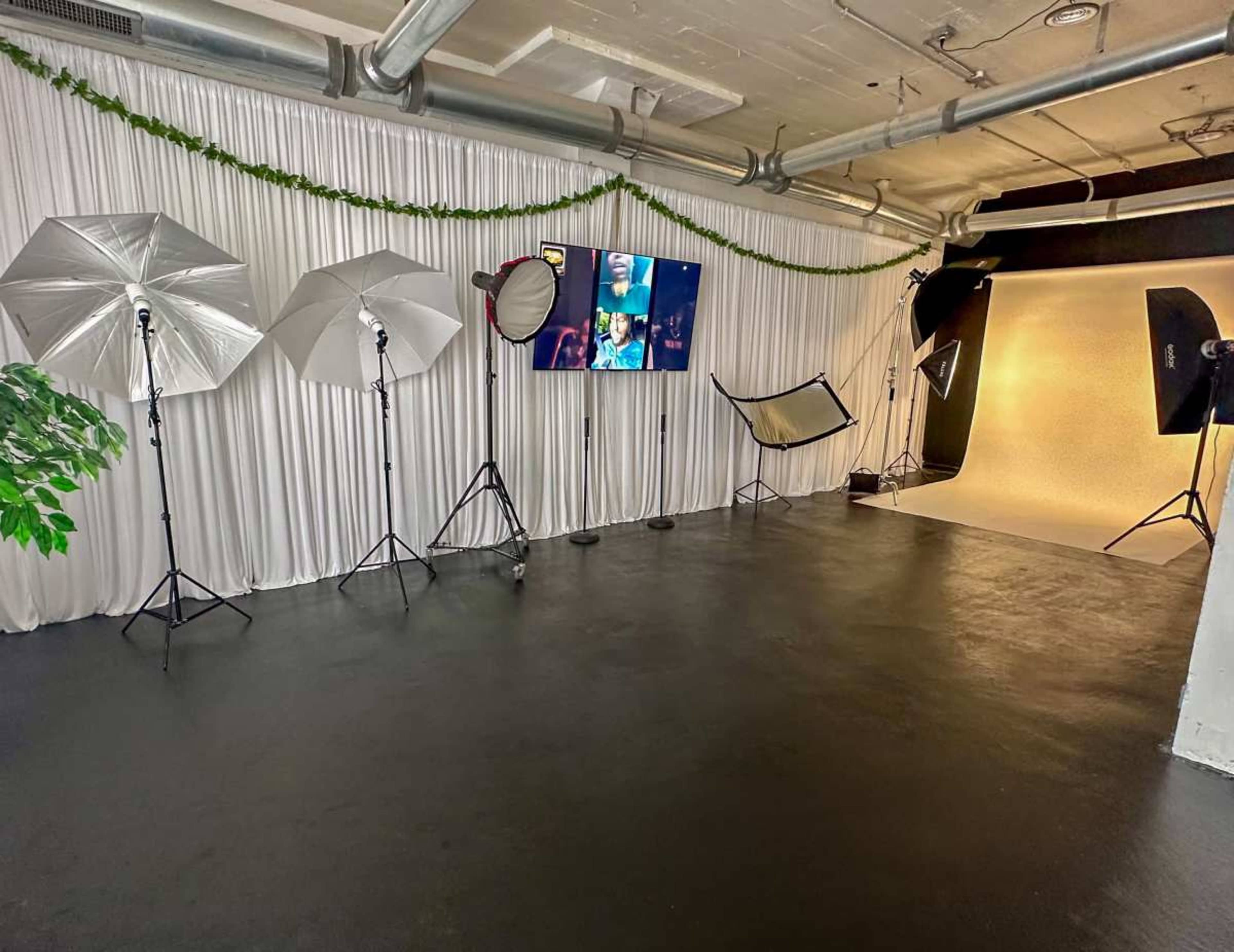 The image shows a photography studio with various lighting equipment set up, including softboxes and reflectors, against a backdrop of white drapery.