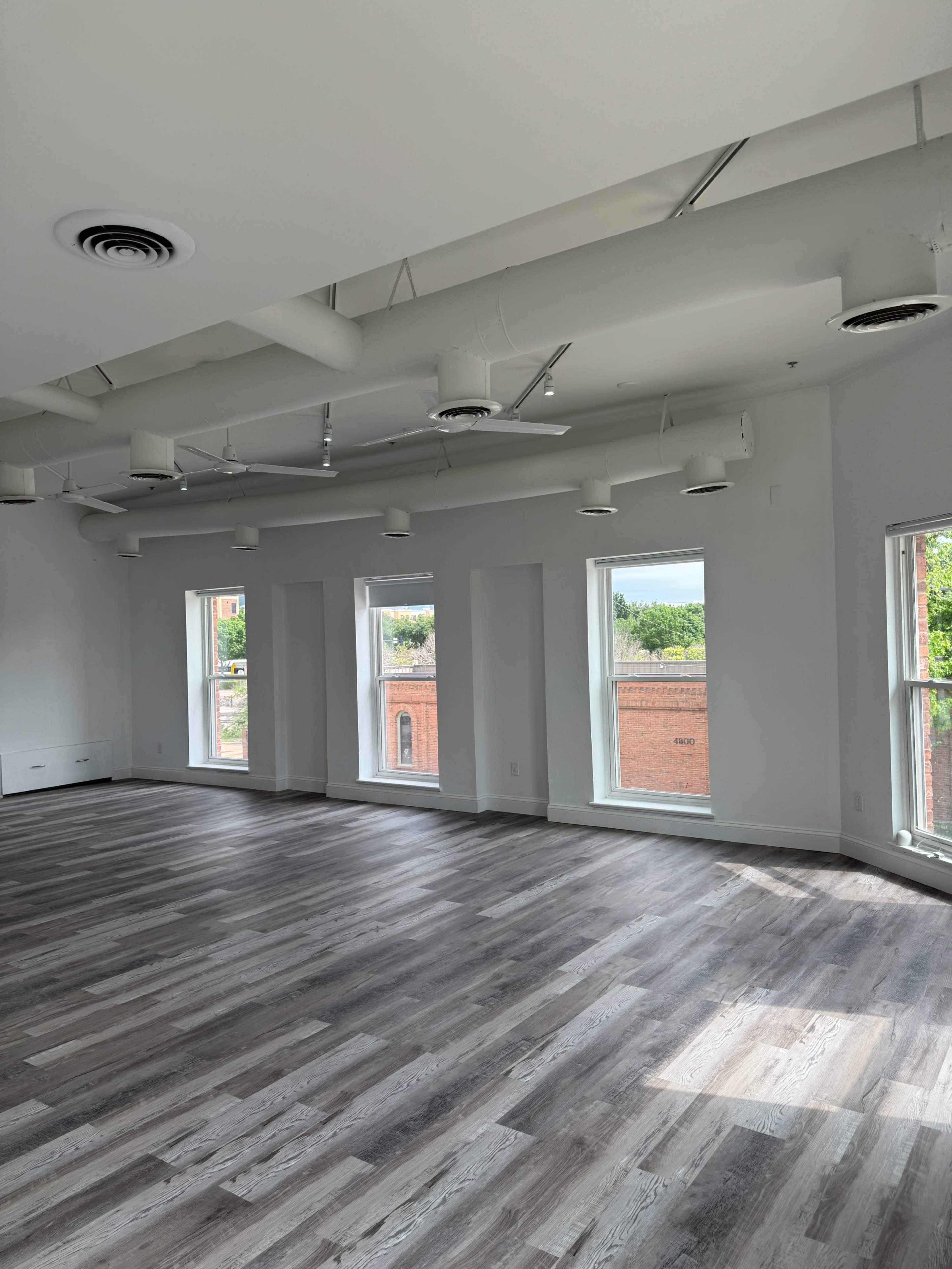 The image shows a spacious, empty room with large windows and wooden flooring.