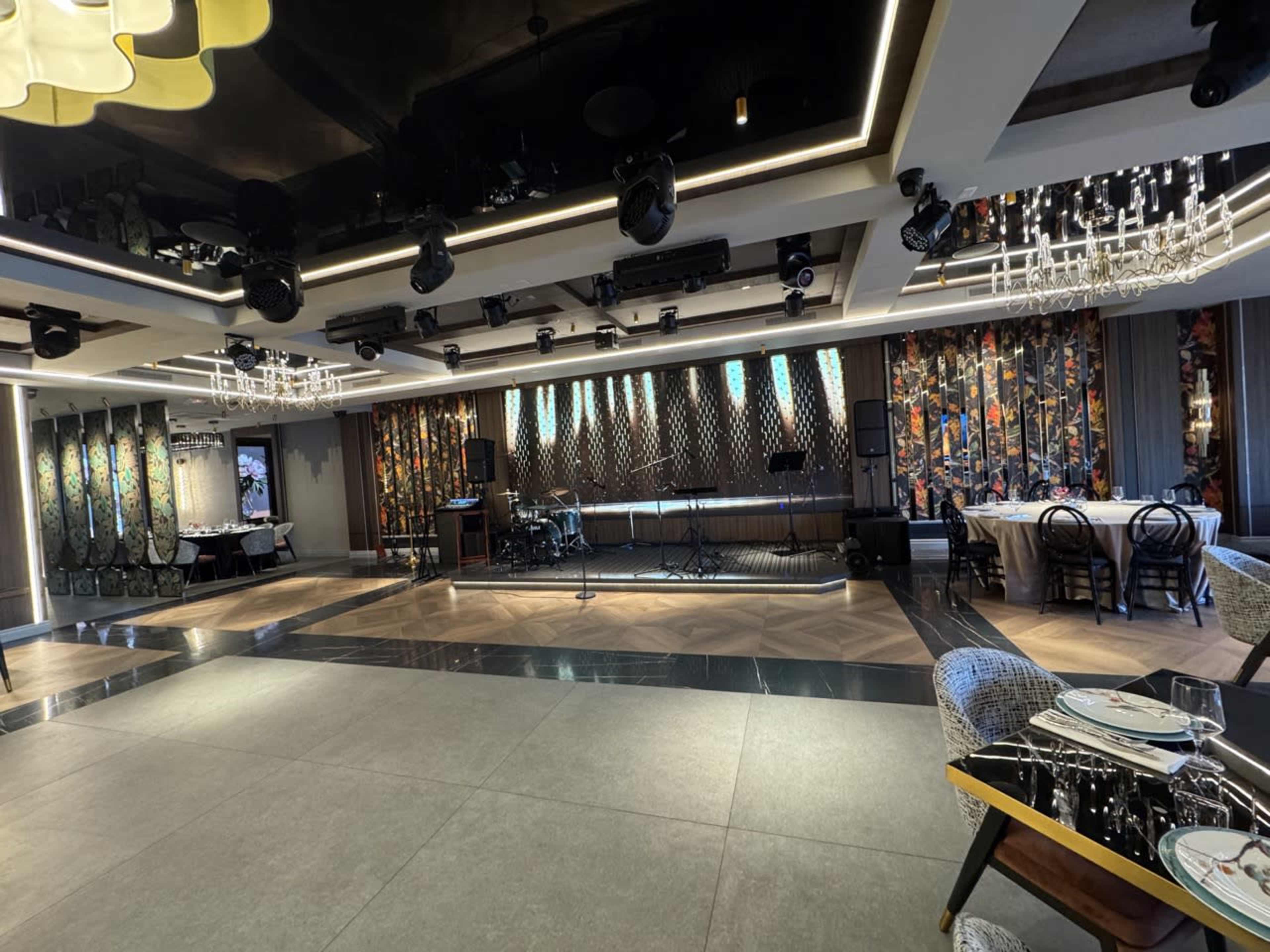 The image shows a modern restaurant interior featuring a stage with musical equipment, colorful wall decorations, and rows of stylish chairs and tables.