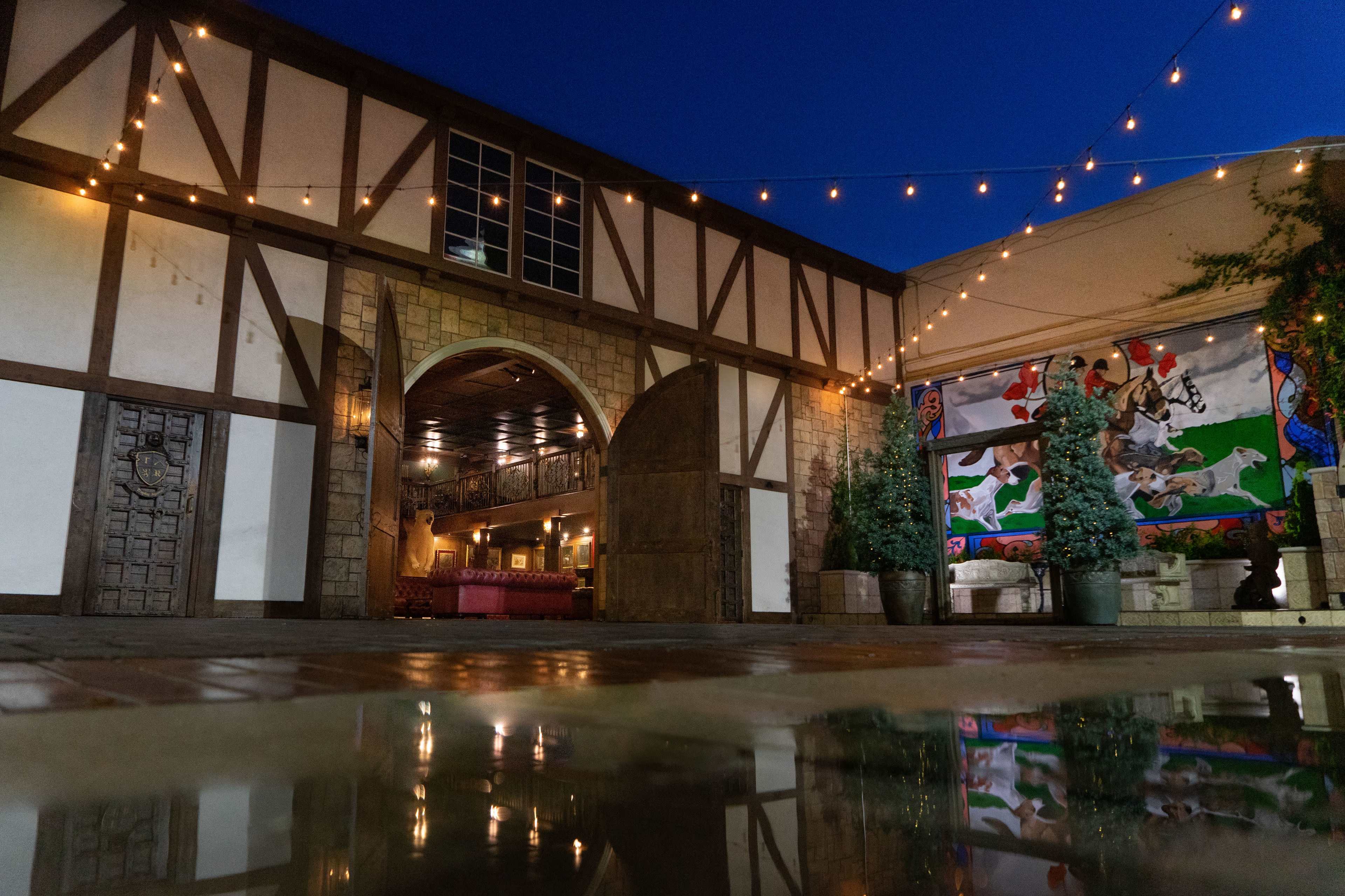 A courtyard with a stone building featuring decorative arches, string lights overhead, and a mural on a nearby wall reflecting in a puddle on the ground.