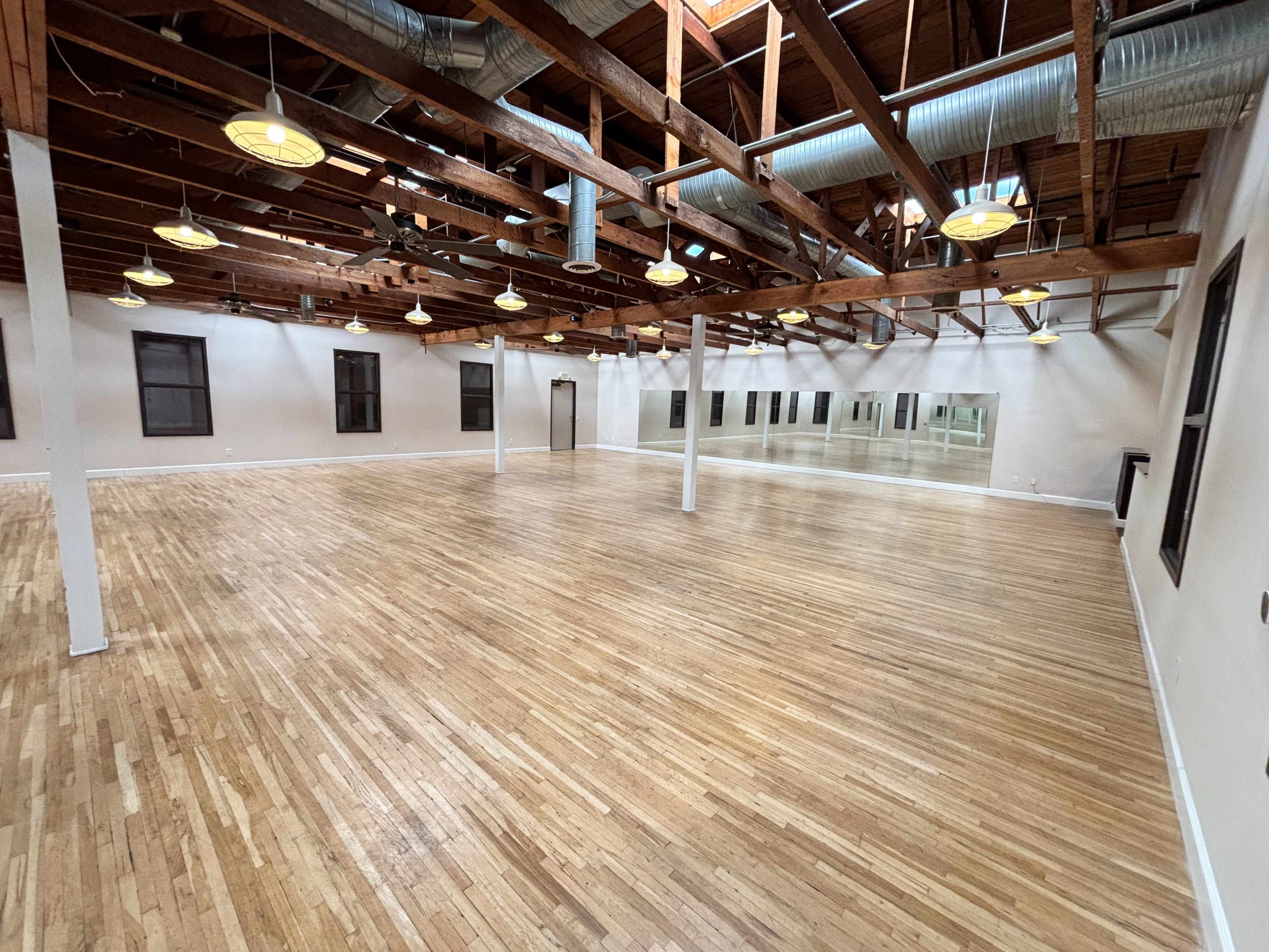 The image shows a spacious, empty room with wooden flooring, exposed wooden beams, and large windows along the walls.
