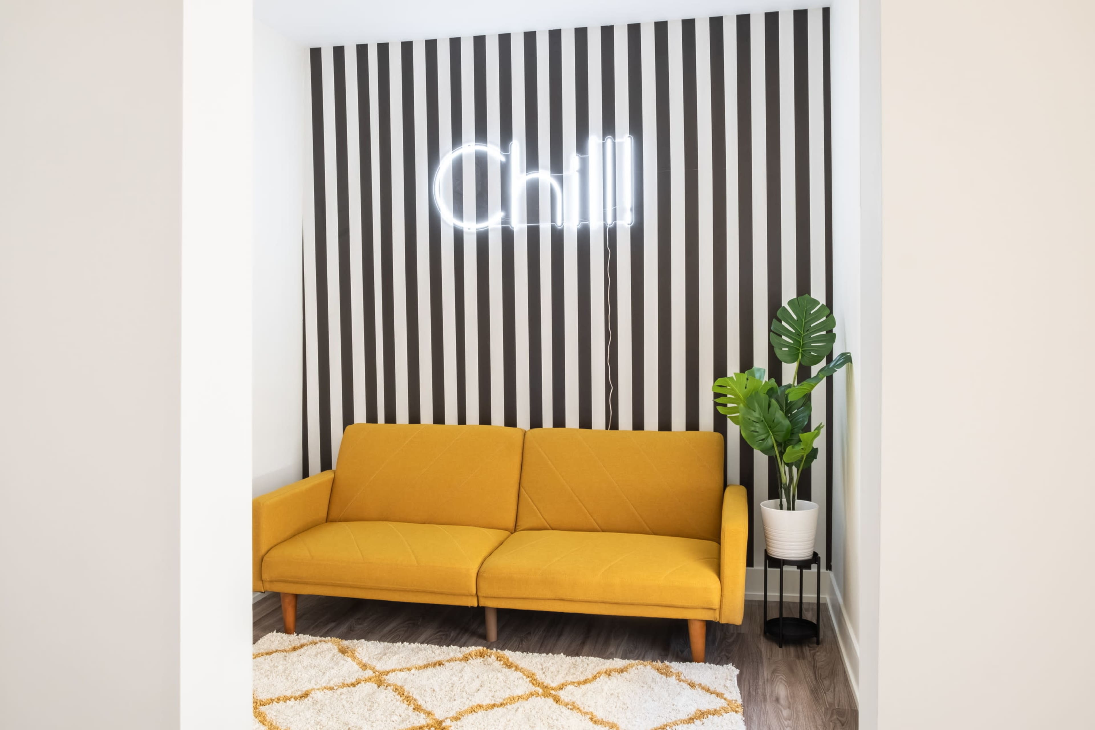 A yellow sofa sits in a room with striped walls, a neon sign that reads "Chill," and a potted plant in the corner.