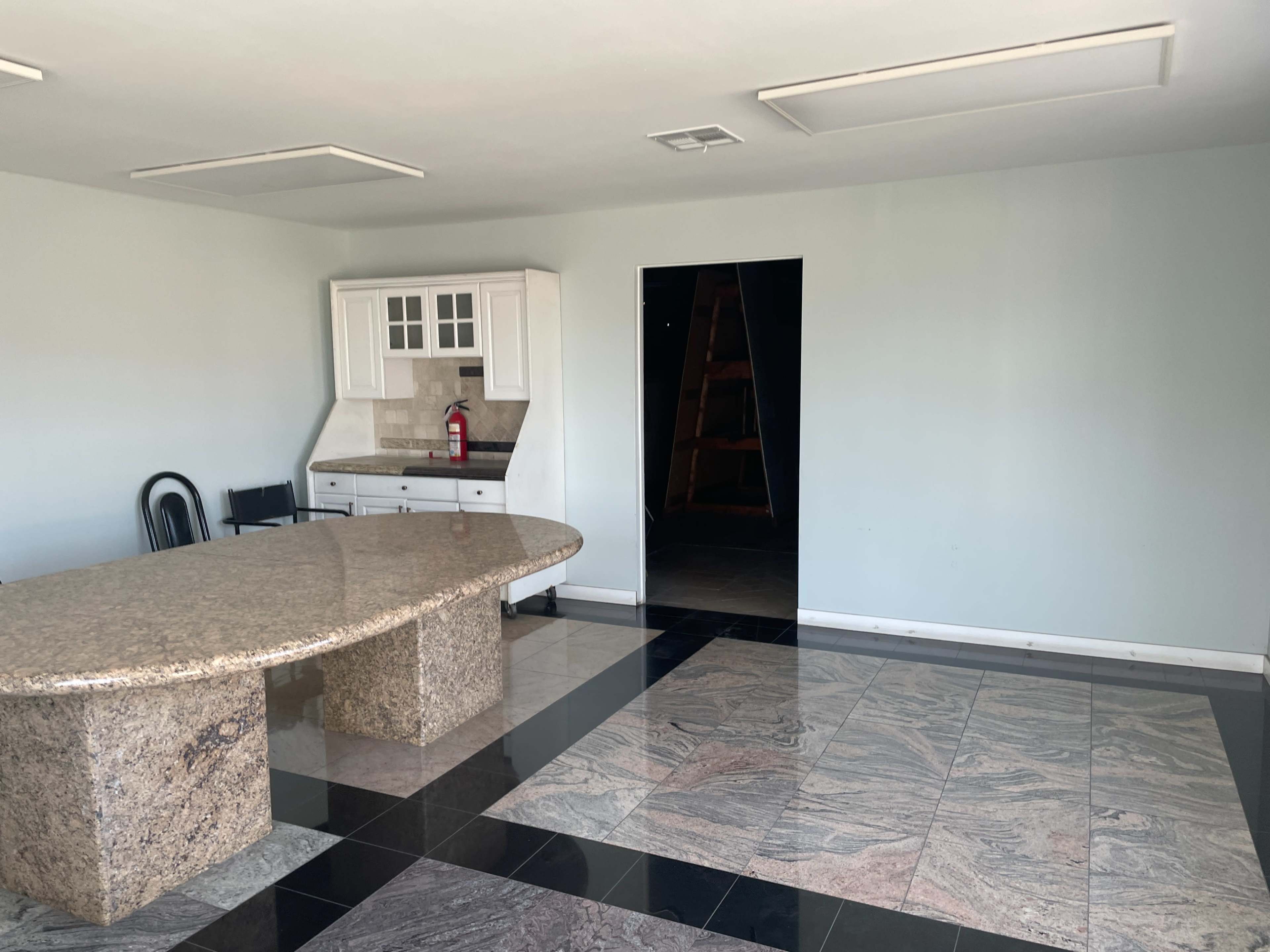 The image shows a spacious room featuring a large granite table with a dark and light tile floor, a white cabinetry set against a pale wall, and an open doorway leading to a darker area.