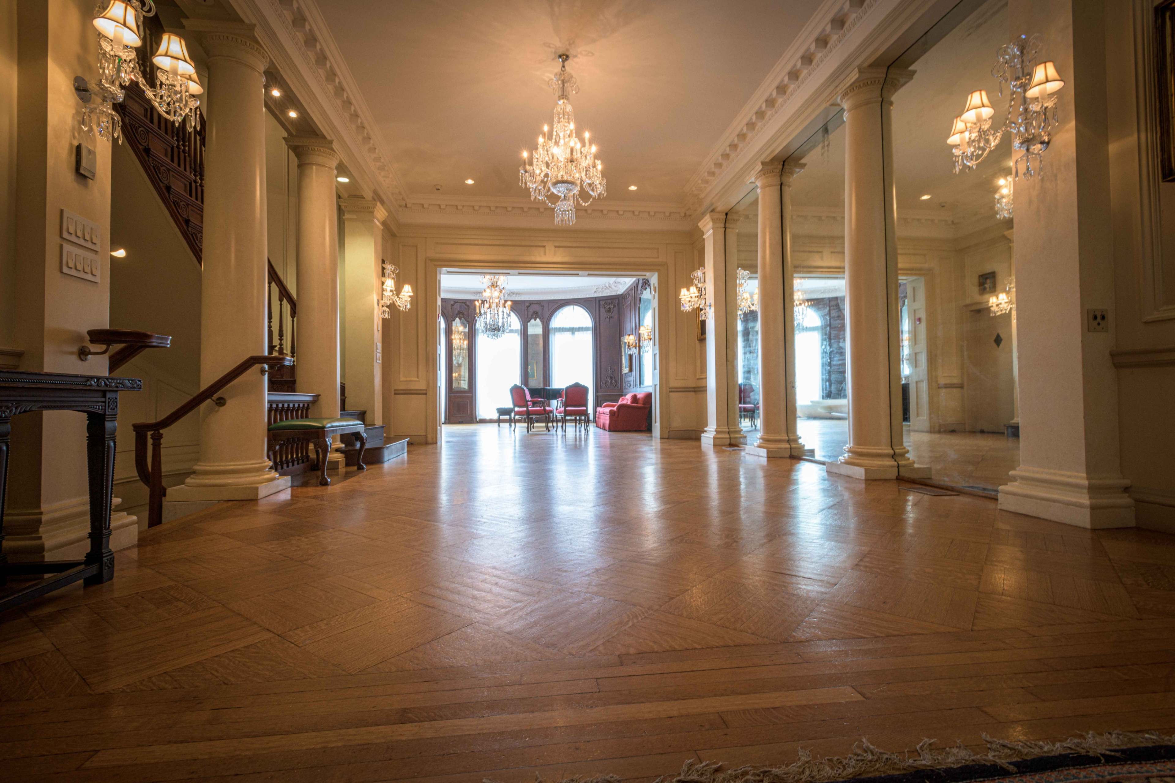 A spacious foyer with polished wooden floors, tall columns, and elegant chandeliers, leading to a living area with red furniture.