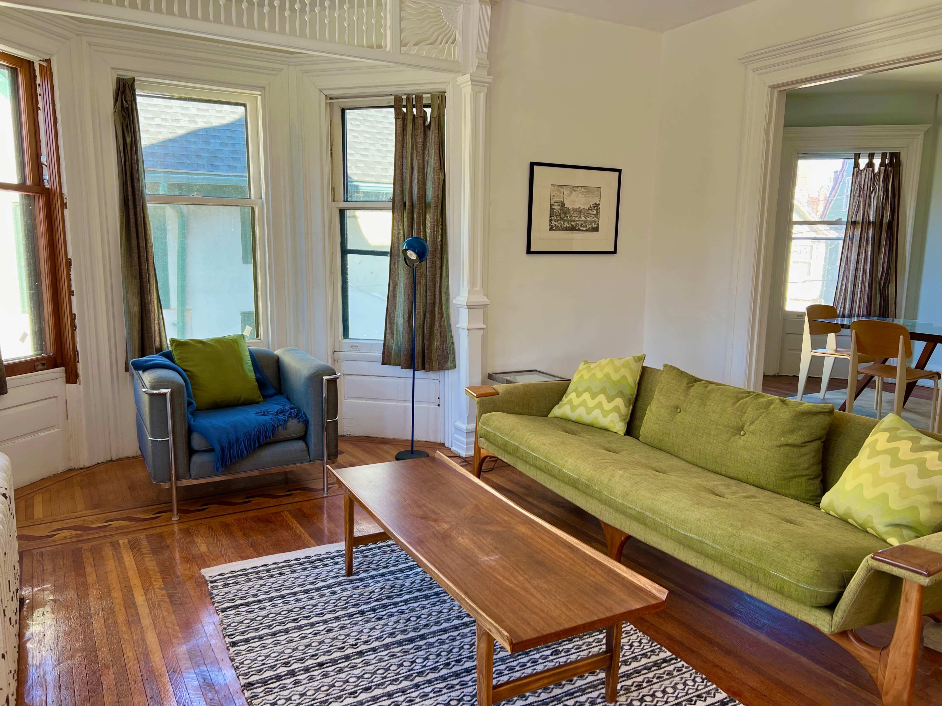 A bright living room with a green sofa, a blue armchair, a wooden coffee table, and two large windows with curtains.