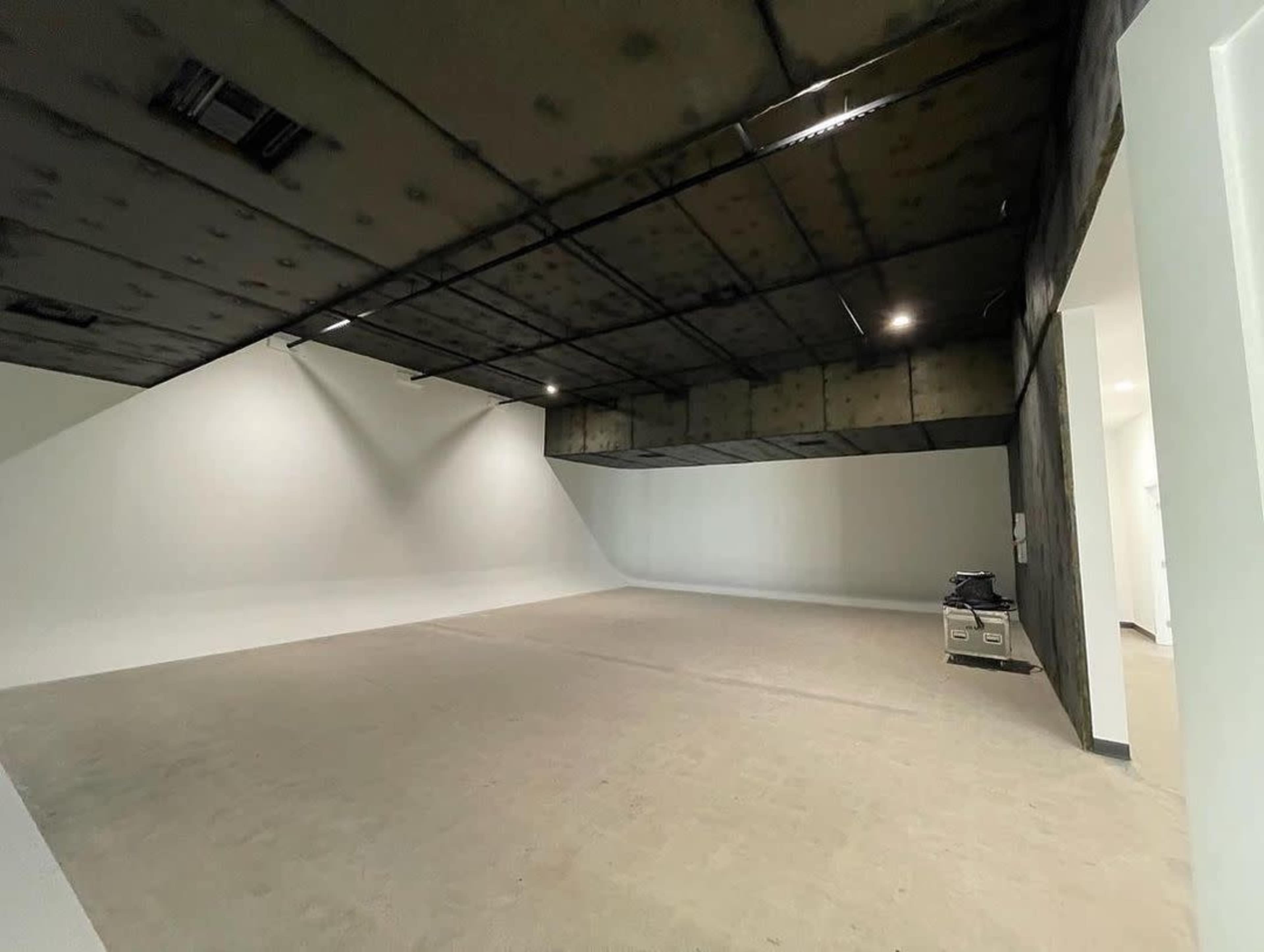 A spacious, empty room with a smooth white wall and a ceiling that is partially blackened, illuminated by overhead lights.