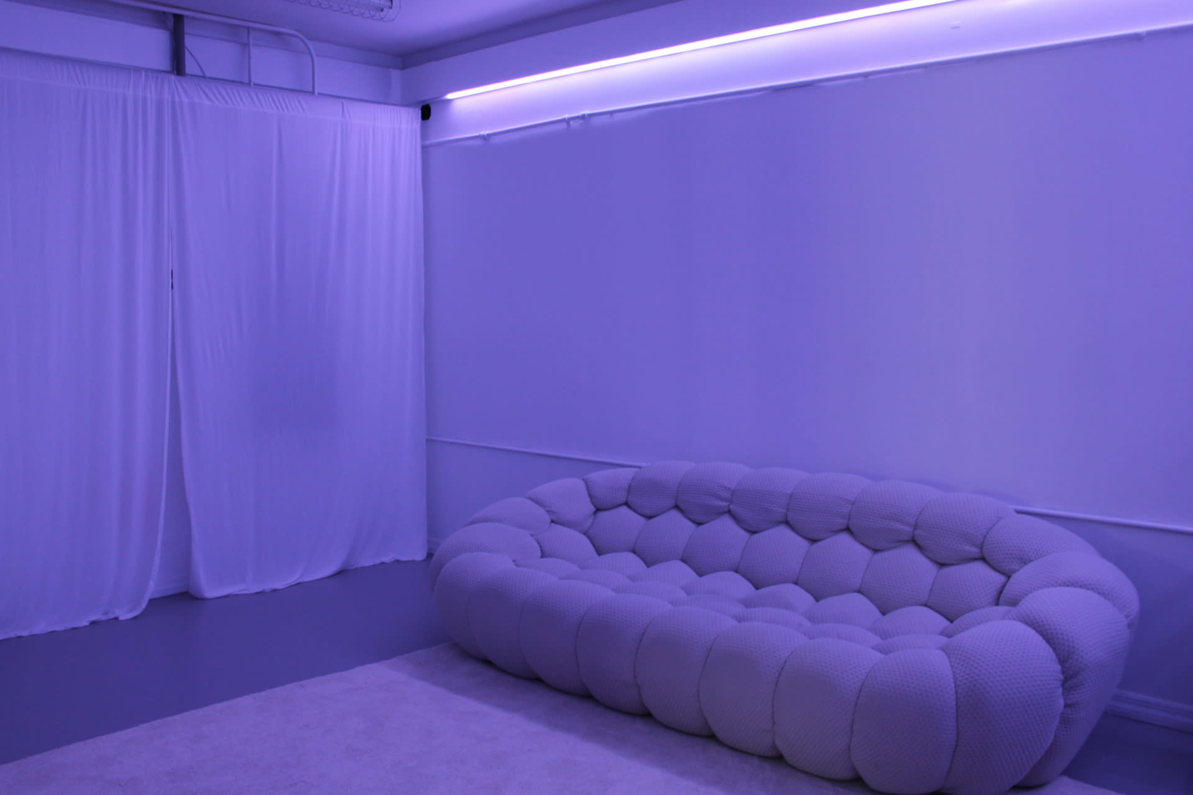 The image shows a modern, plush, gray couch against a softly lit purple wall with draped white curtains.