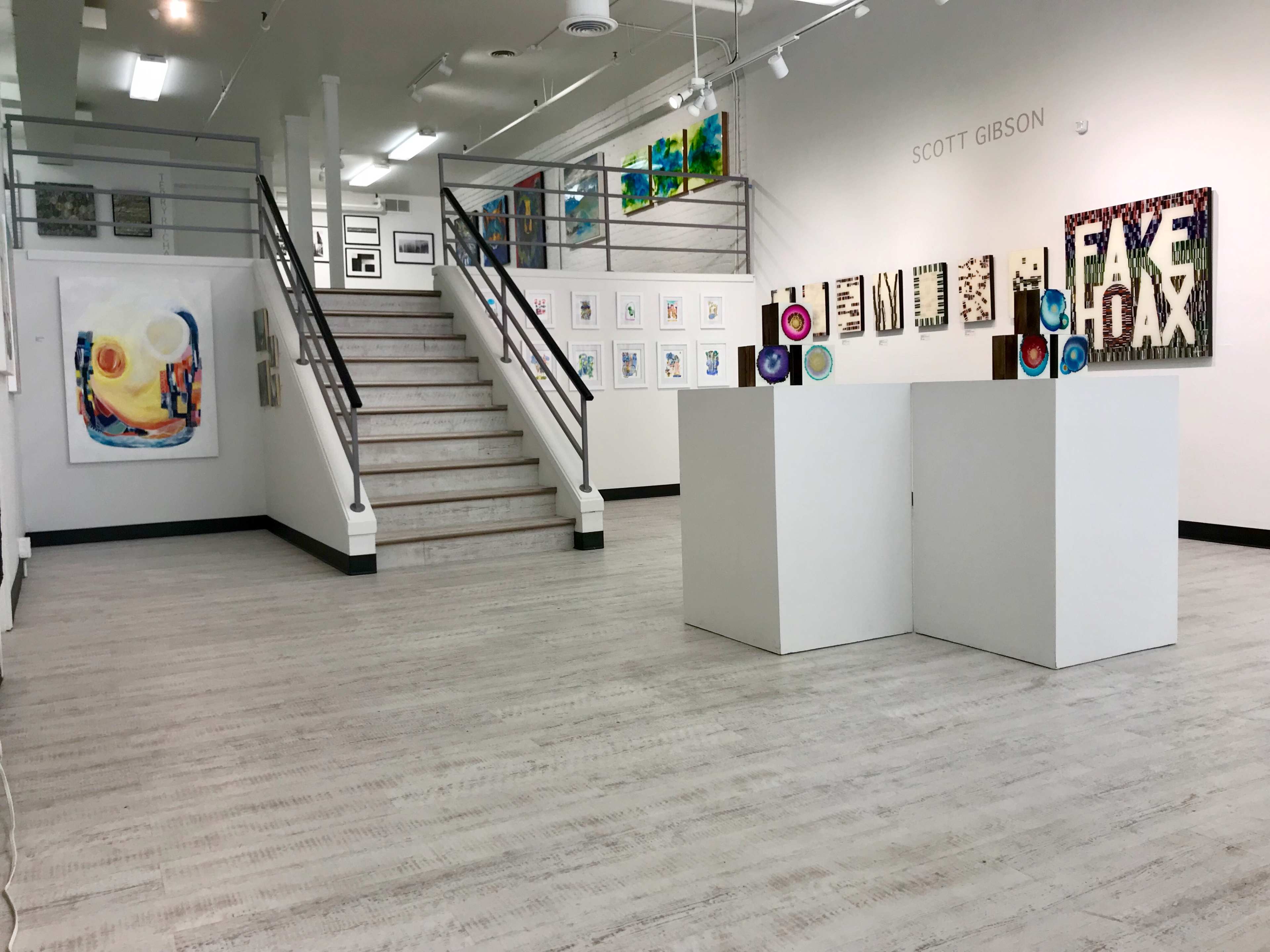 A modern art gallery with white walls, wooden floors, and a staircase leading to an upper level, displaying various artworks in frames and on pedestals.