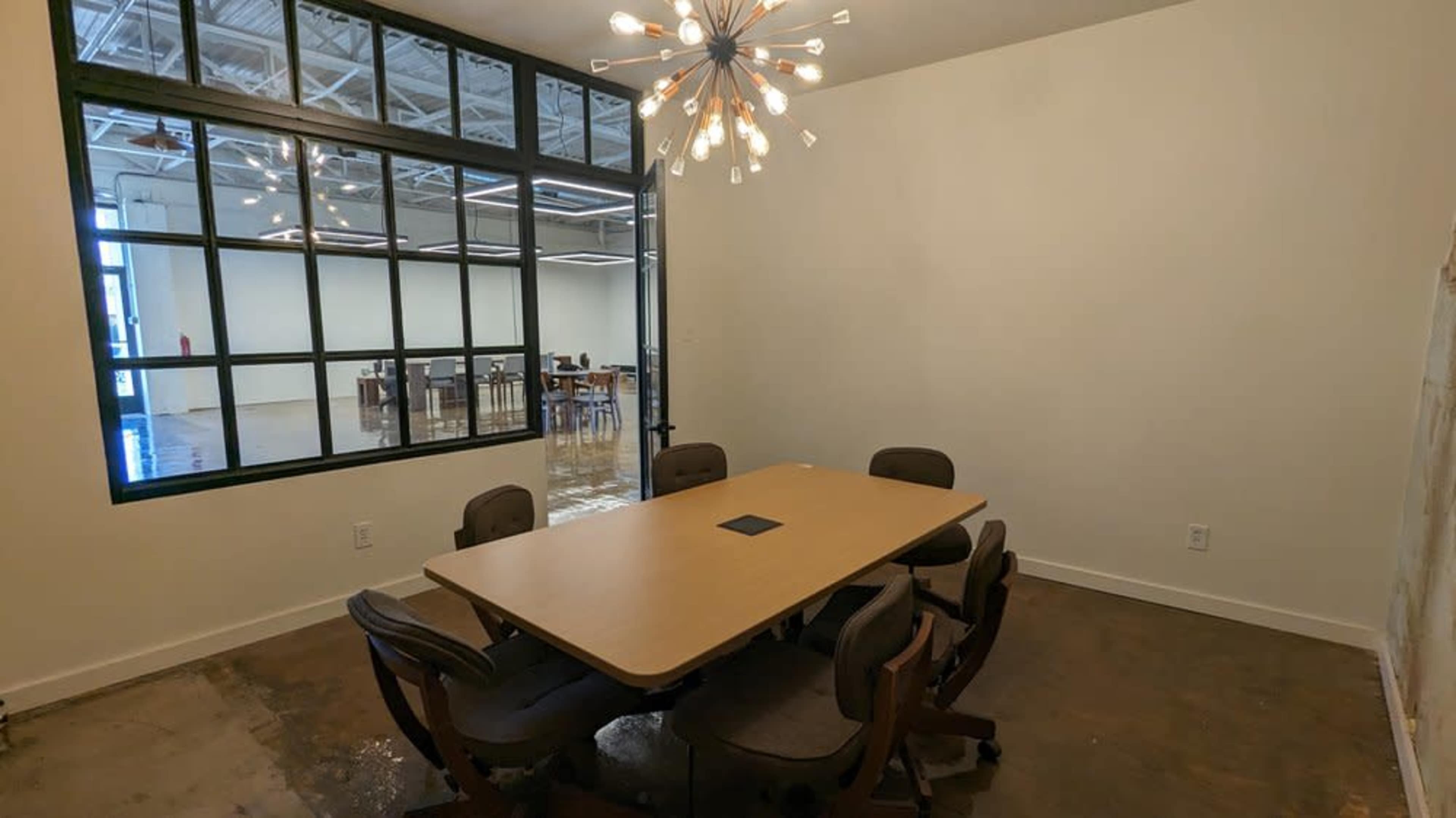 A conference room features a large table surrounded by eight chairs, with a modern light fixture overhead and a window providing a view of another workspace.