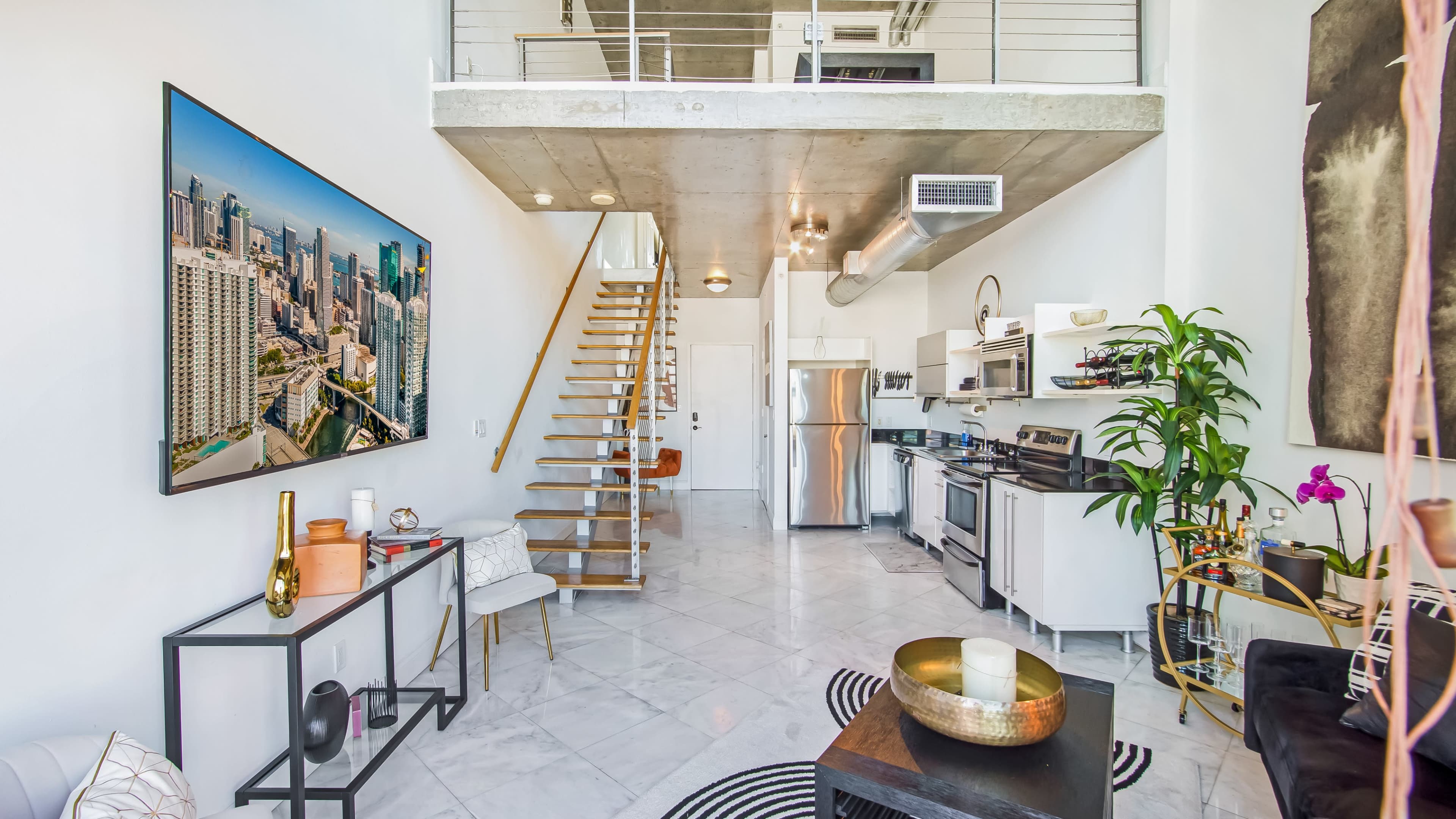 A modern, open-concept living space features a staircase leading to a loft, a kitchen with stainless steel appliances, and large windows showcasing a city view.