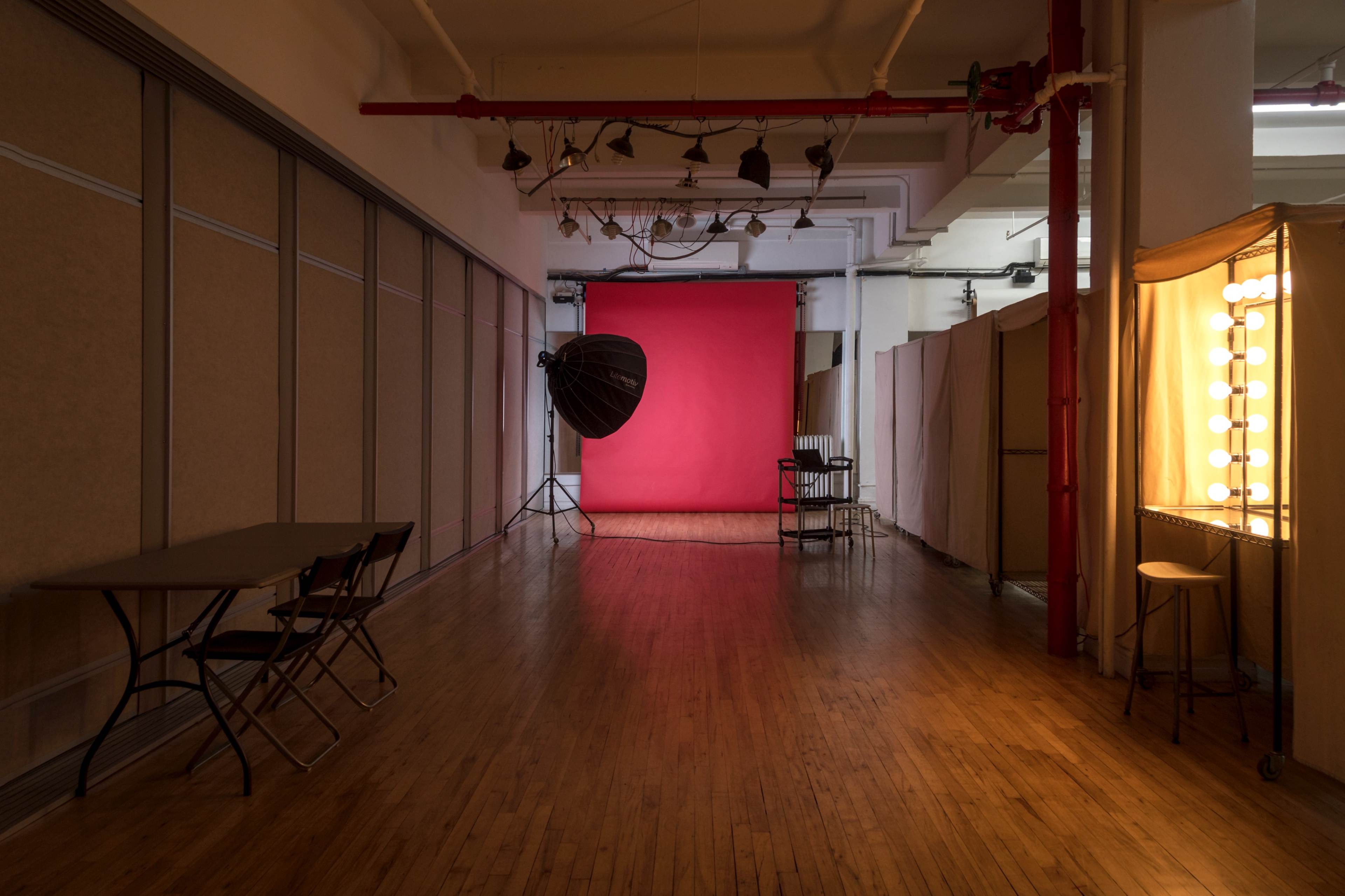 The image shows a photography studio with a red backdrop, softbox light, and a row of dressing room mirrors along one side.
