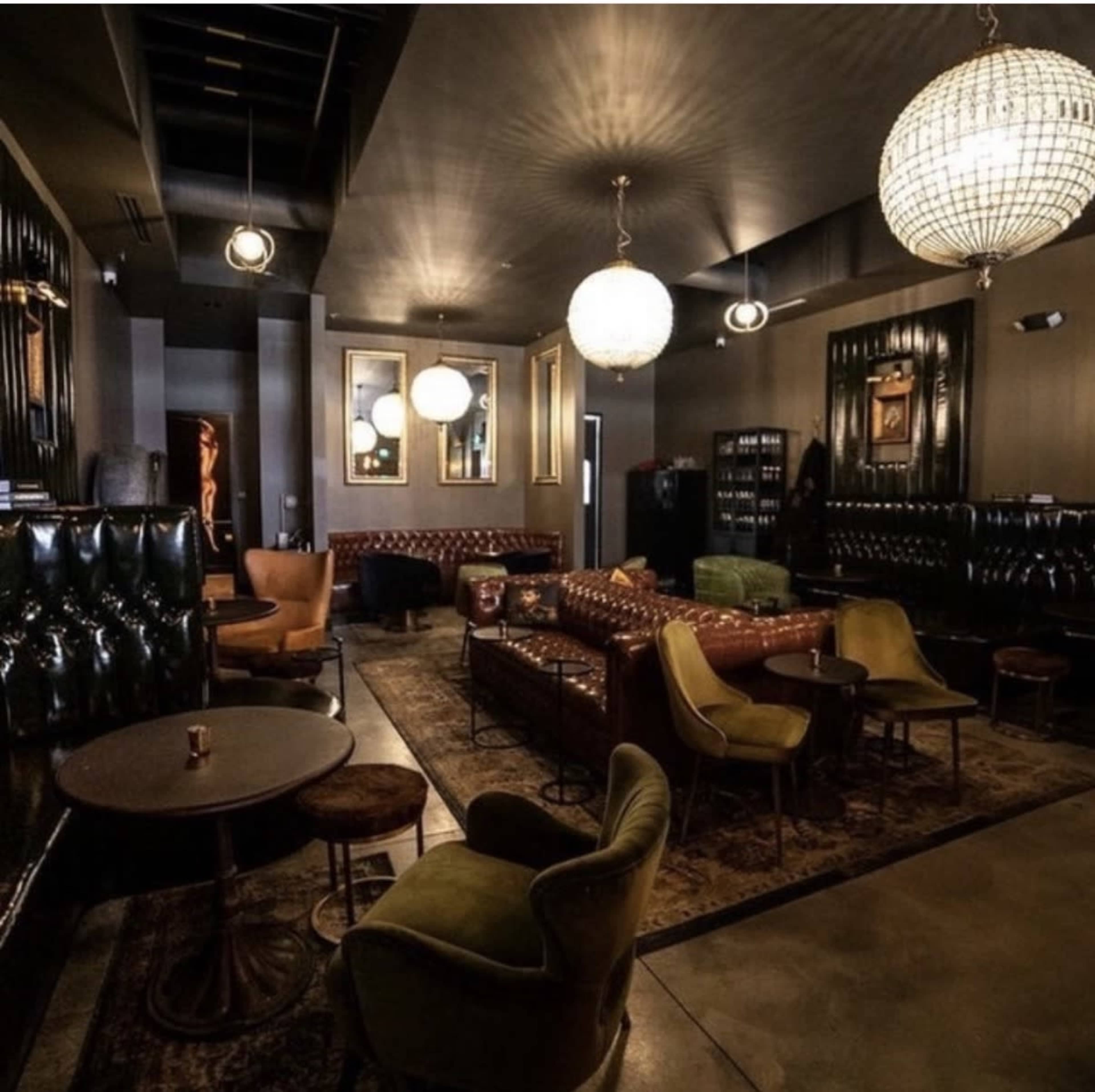 The image depicts a stylish lounge area featuring dark walls, plush seating, and decorative chandeliers.