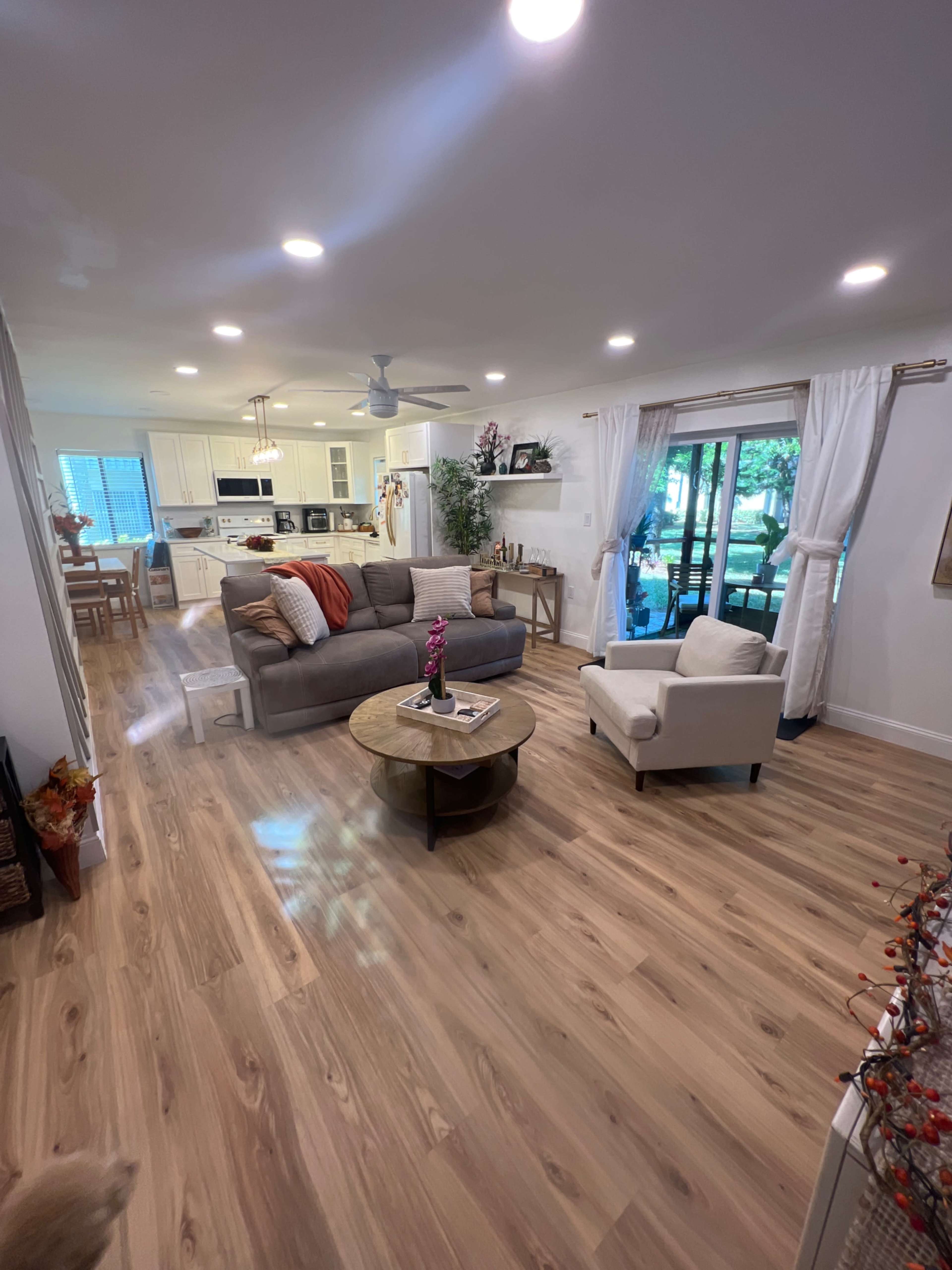 The image shows a modern living room with a gray sofa and a white armchair, a round coffee table, and large windows providing natural light.