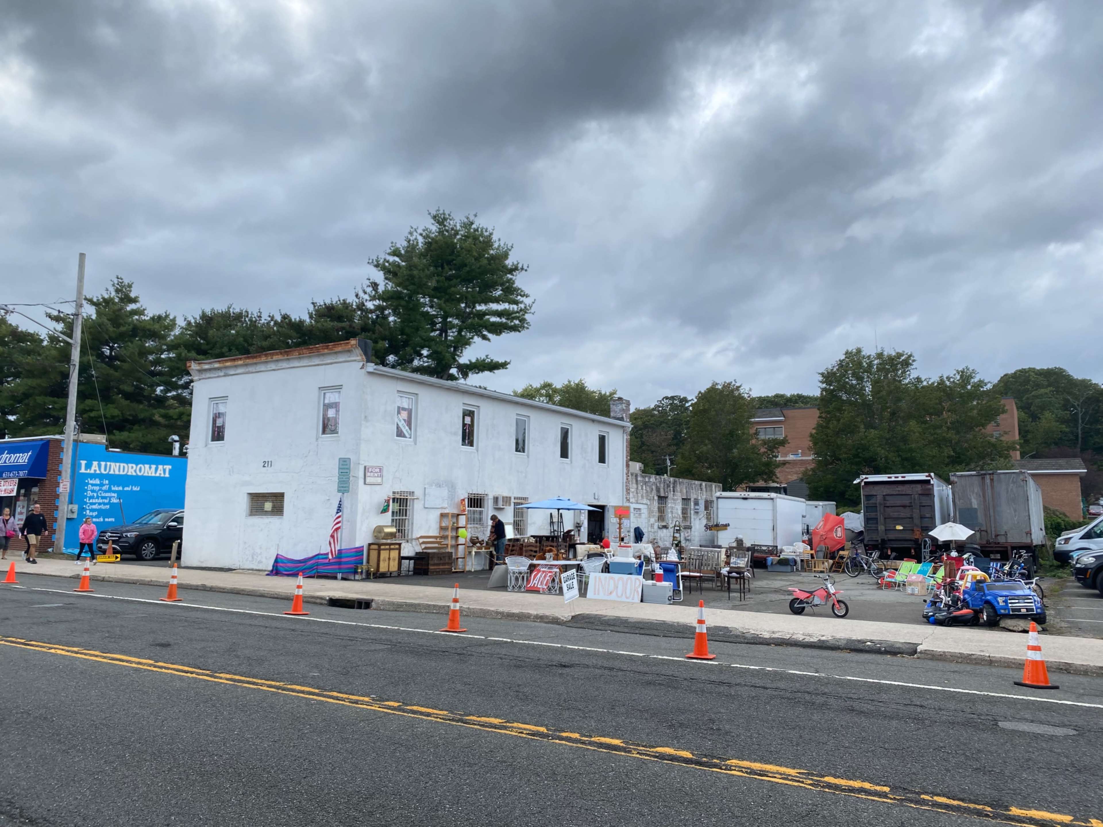 A long, white building with several windows sits beside a road, surrounded by various items for sale and traffic cones blocking parts of the street.