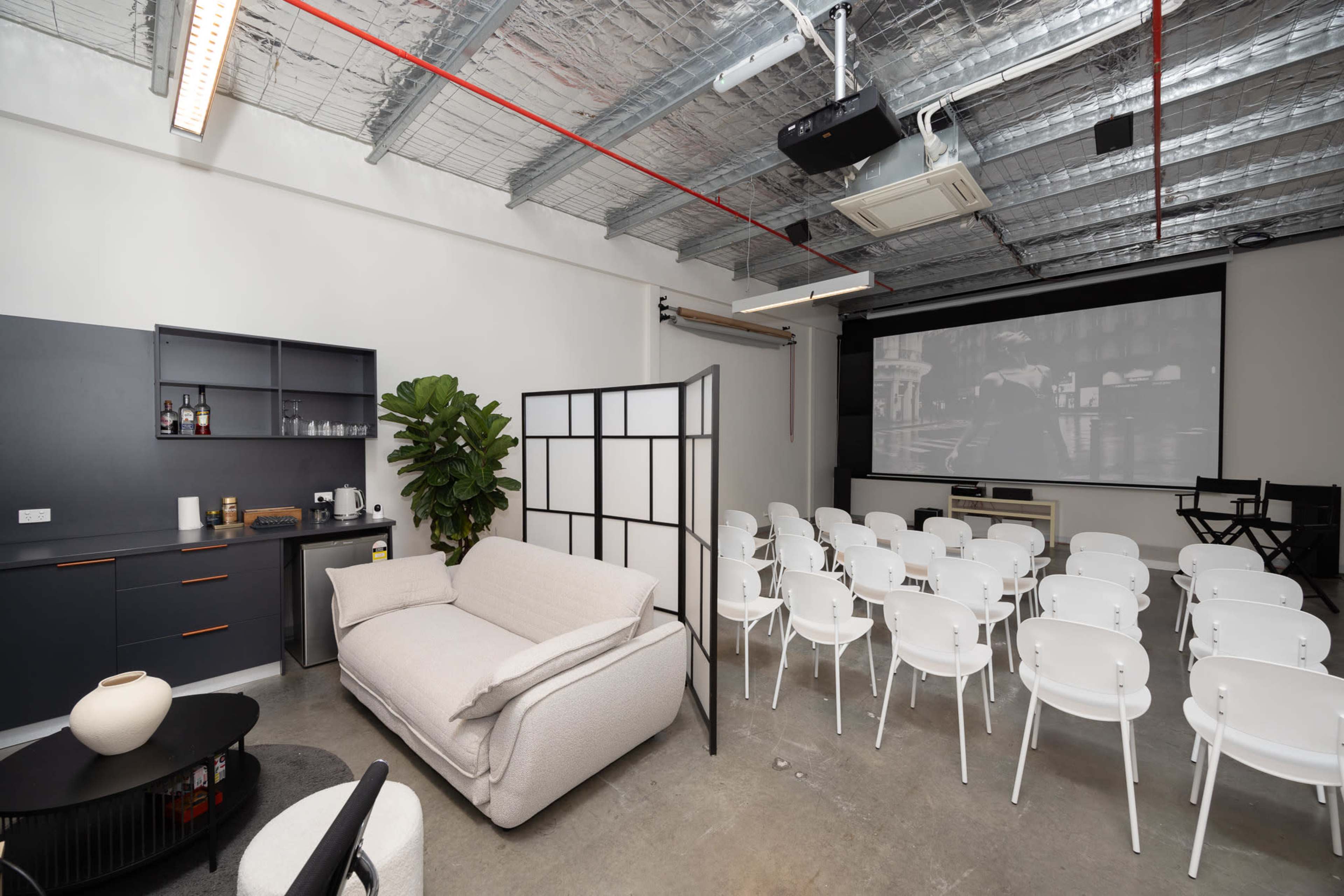 The image shows a modern room equipped with a projector screen, white chairs arranged in rows, a sofa, and a kitchenette area.