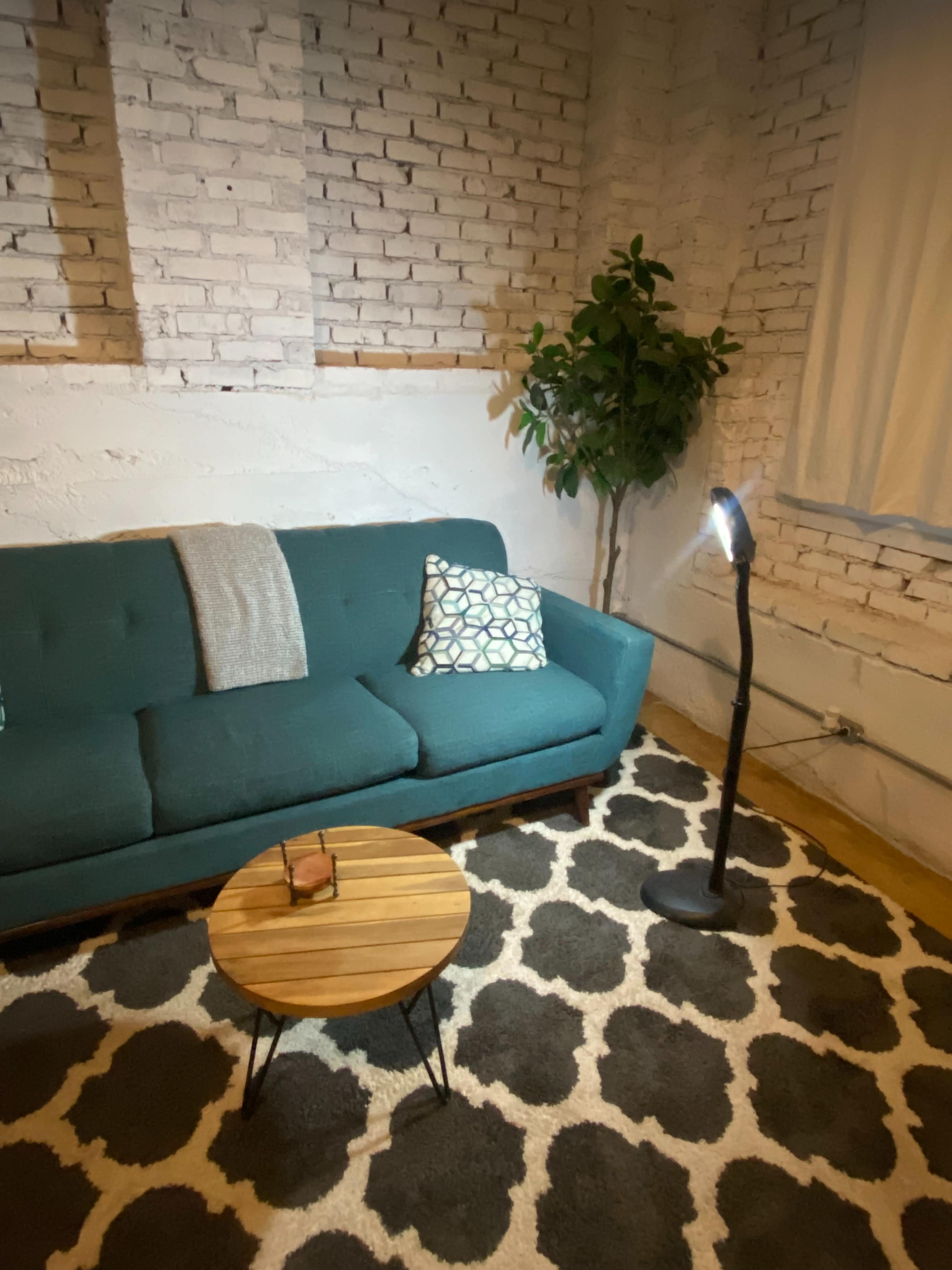 The image shows a cozy living room with a teal couch, a small wooden coffee table, a standing lamp, and a potted plant against a white brick wall.