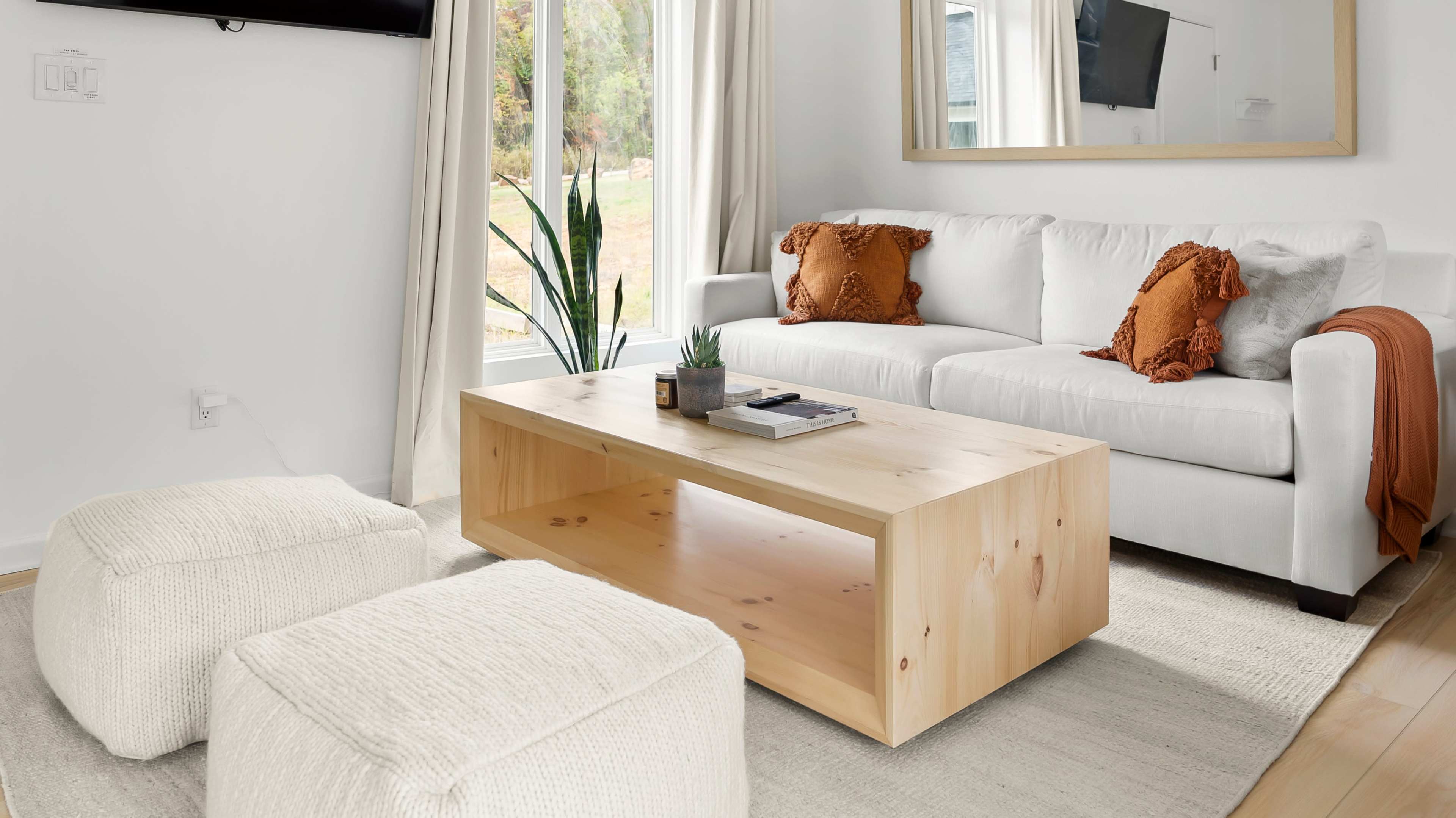 The image features a modern living room with a white sofa adorned with orange cushions, a wooden coffee table, and two round, textured ottomans.