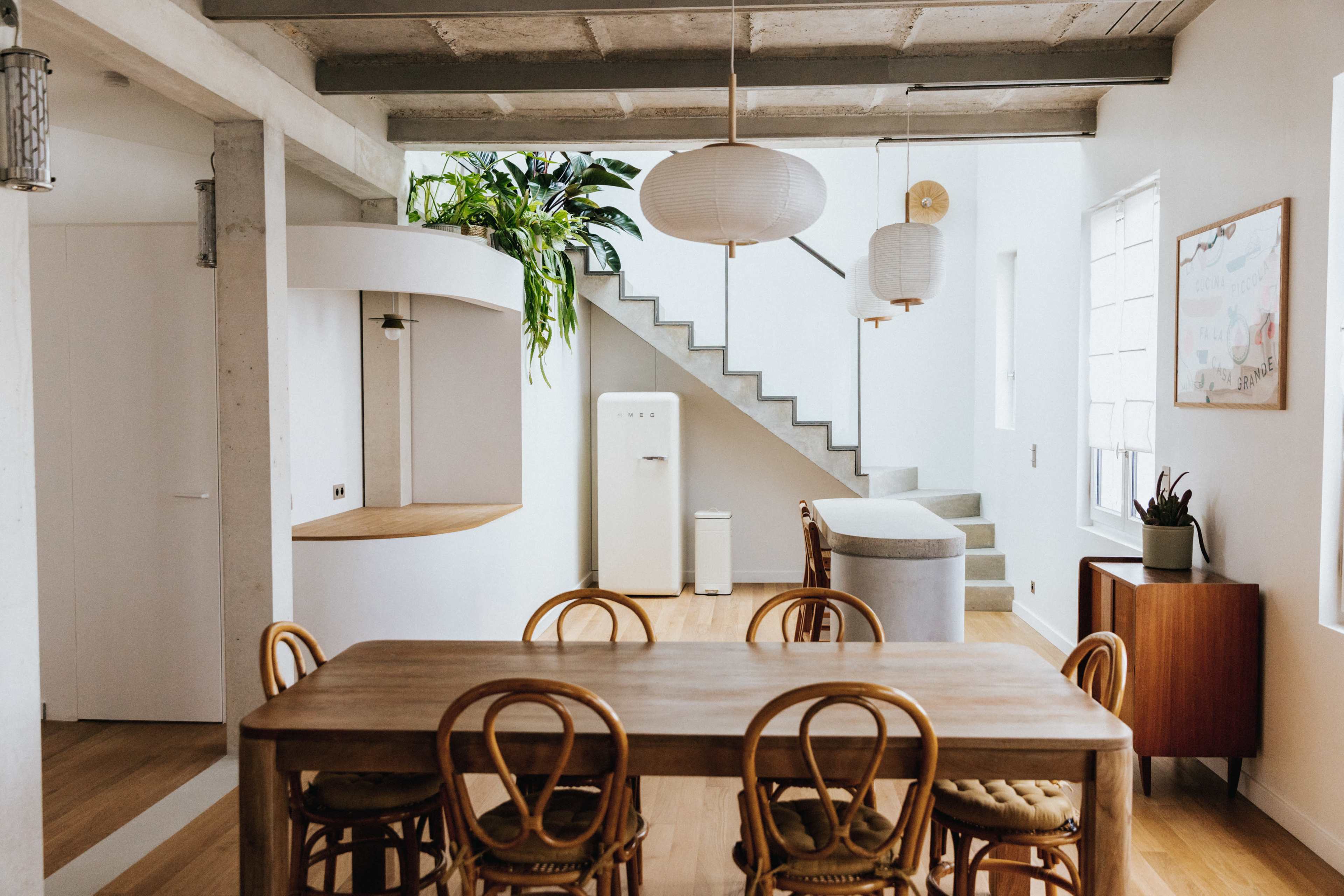 A modern dining area features a wooden table surrounded by chairs, with a staircase in the background leading to an upper level, illuminated by hanging pendant lights.