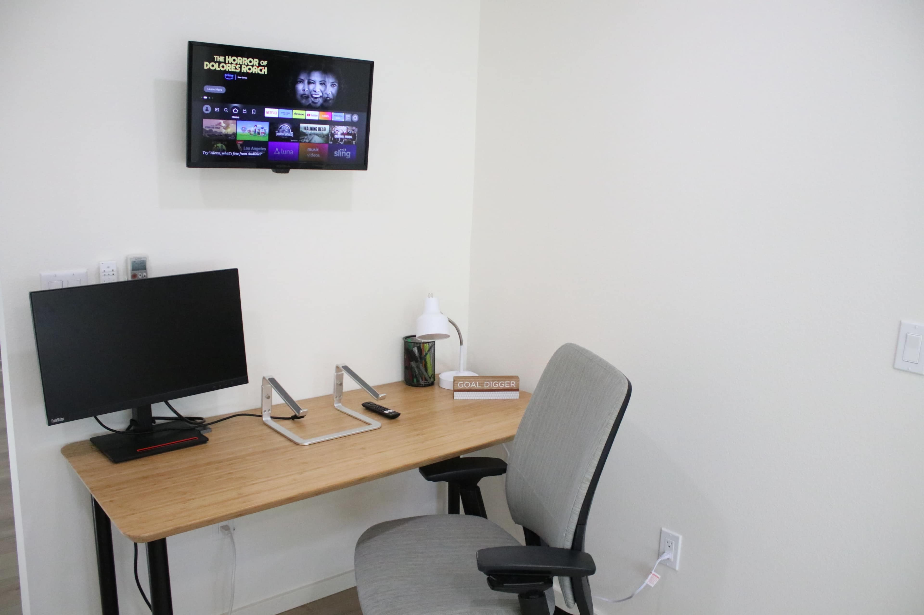 A wooden desk with a computer monitor and a chair is positioned under a wall-mounted television in a minimalistic room.