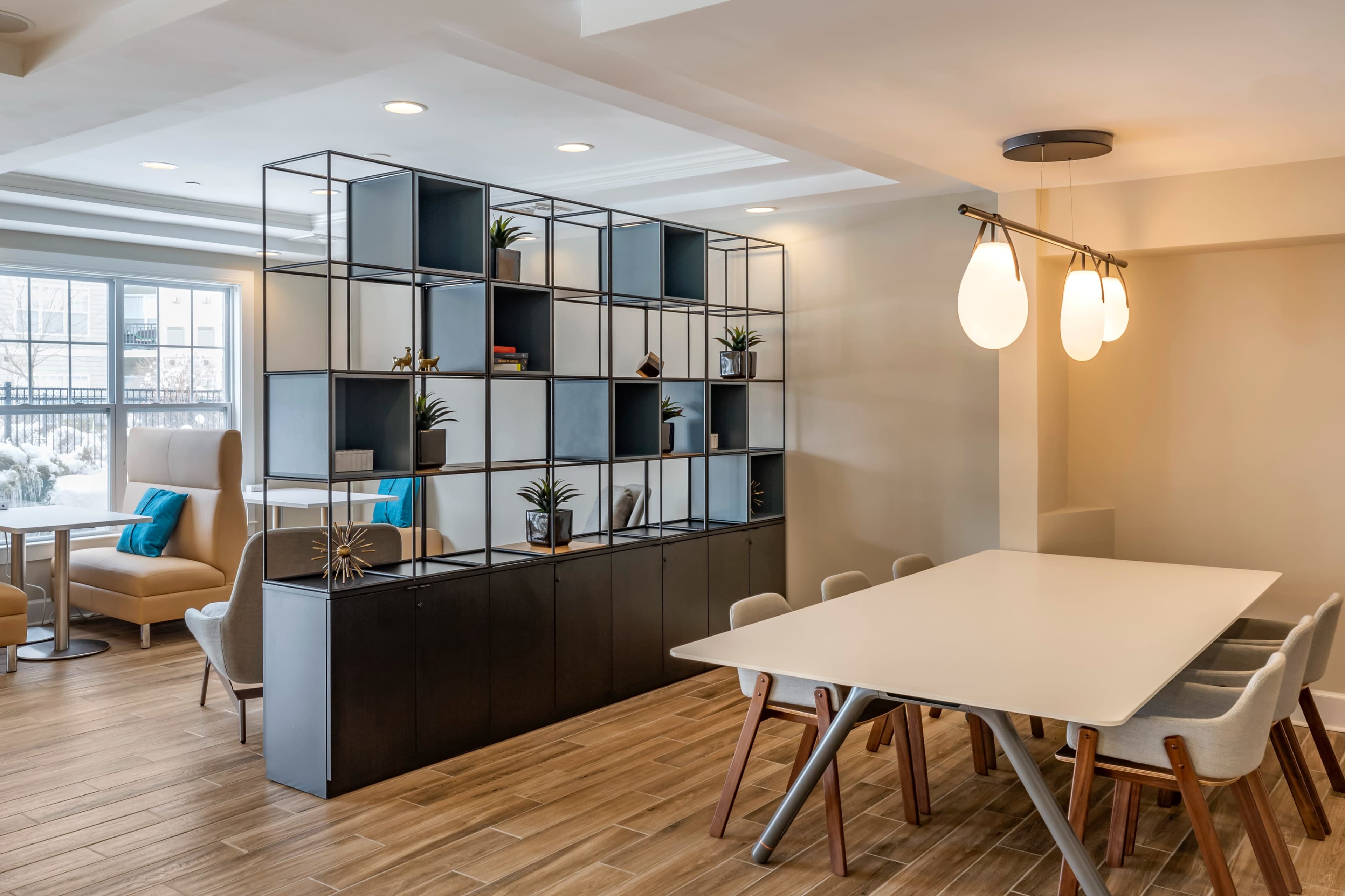 A modern dining area with a long table, light-colored chairs, and a geometric shelving unit filled with plants and decorative items.