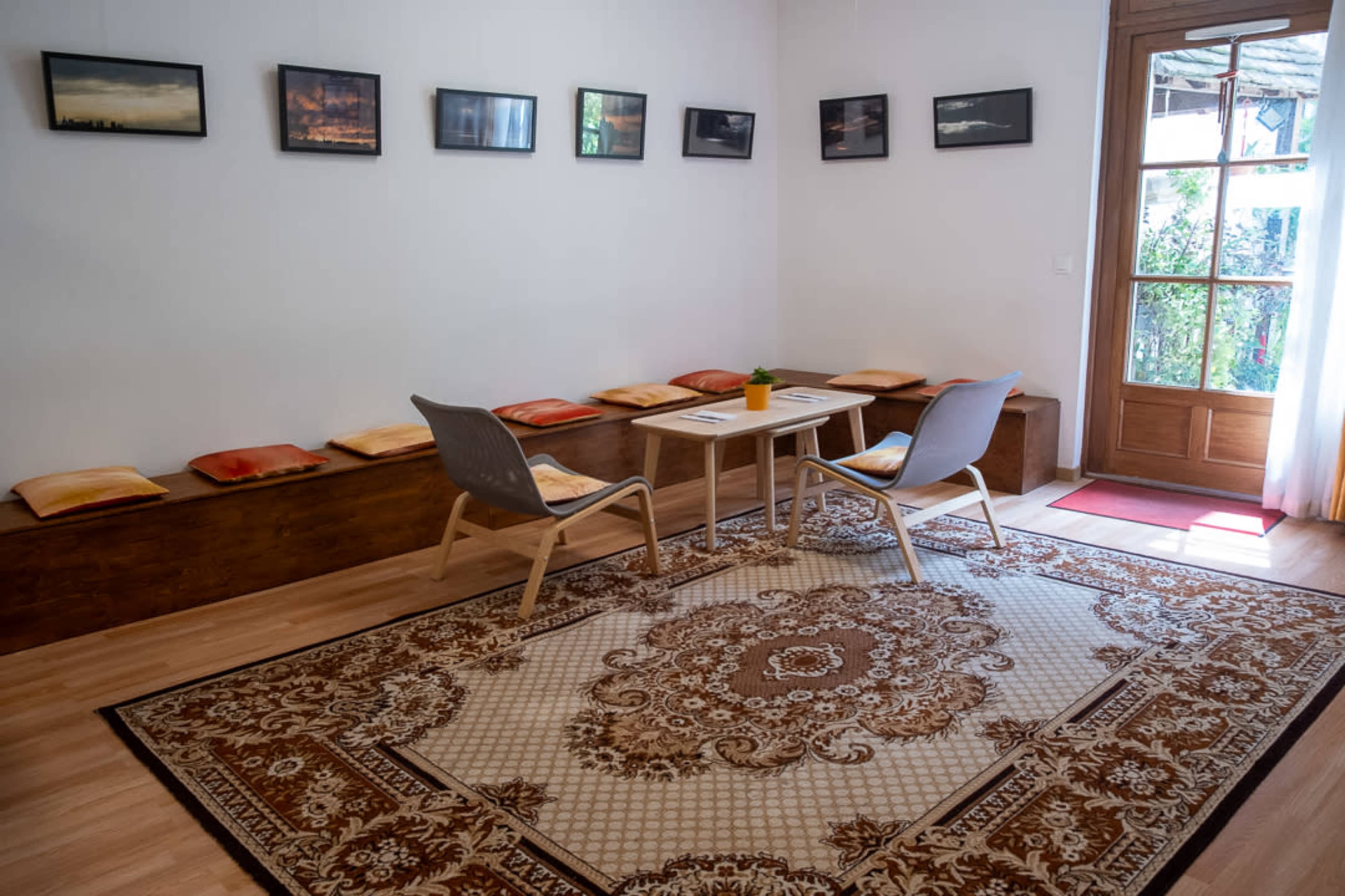 A cozy room features a wooden bench with cushions, a small table, two chairs, and framed photographs hanging on the walls, along with a door leading outside.