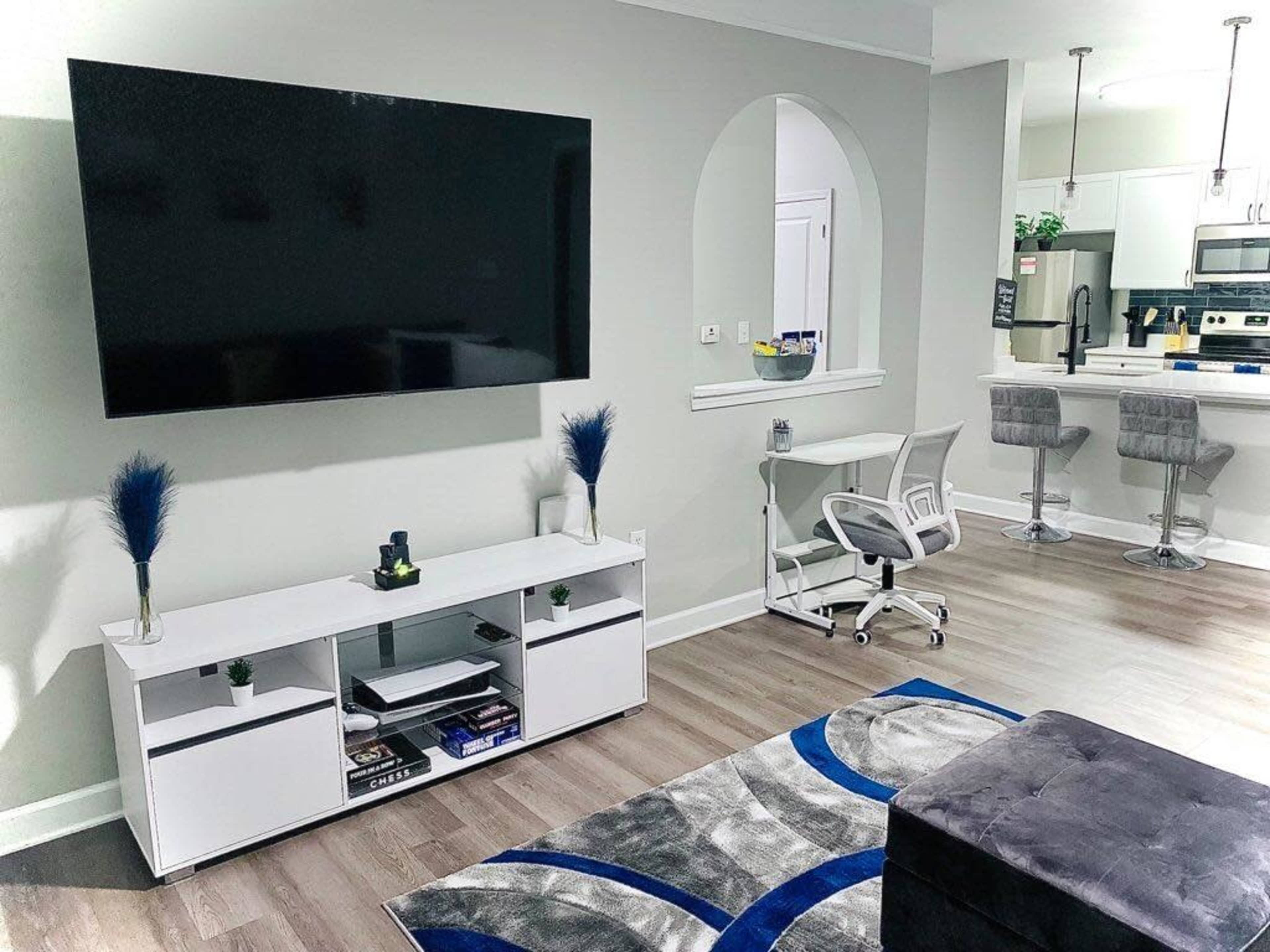 A modern living room with a large flat-screen television mounted on the wall, a white entertainment center, a desk with a chair, and a small kitchen area visible in the background.